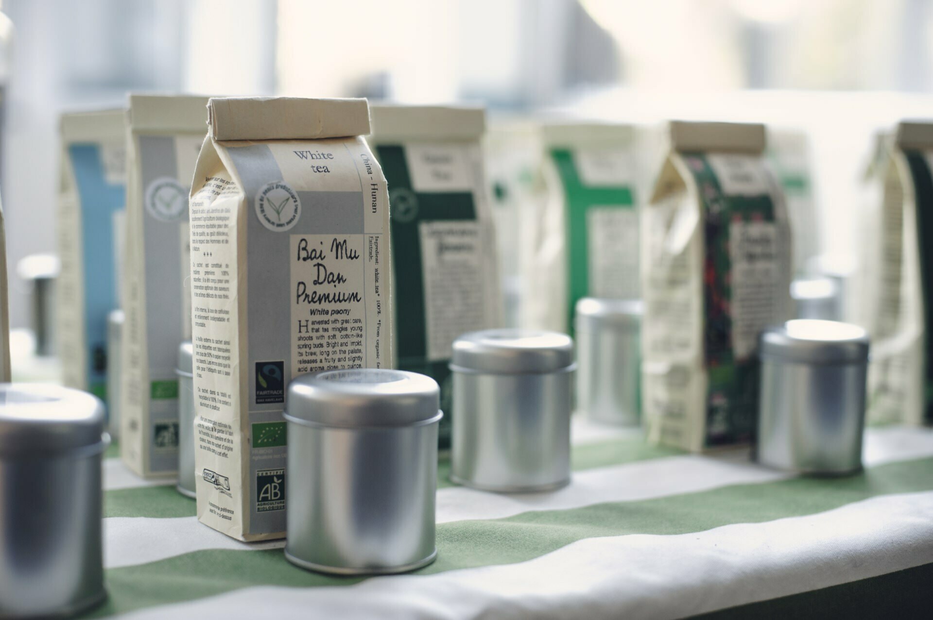 A collection of tea packs and cylindrical metal cans is displayed on a table with a green and white striped tablecloth, referring to a food fair. The packages have different labels and are arranged in rows, with the "Bai Mu Dan Premium" label in the foreground. 