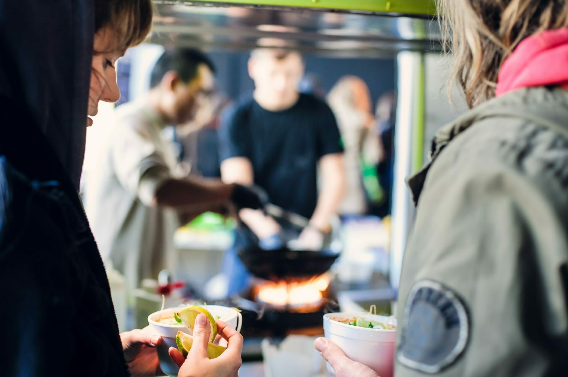 People gather around a street food vendor at a food market. Two people in the foreground hold bowls of food while the vendor in the background, dressed in a black t-shirt and gloves, prepares a dish on a large wok over an open fire. 