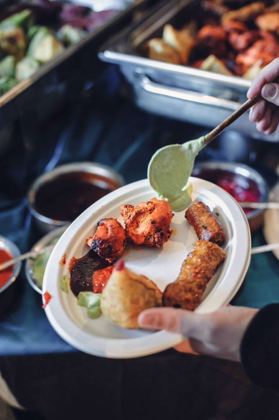 Osobie trzymającej biały papierowy talerz z różnymi indyjskimi przystawkami, w tym samosą, tikką i sajgonką, podaje się łyżką zielony chutney. W tle tej sceny targów kulinarnych na tacach widać inne indyjskie potrawy i sosy.