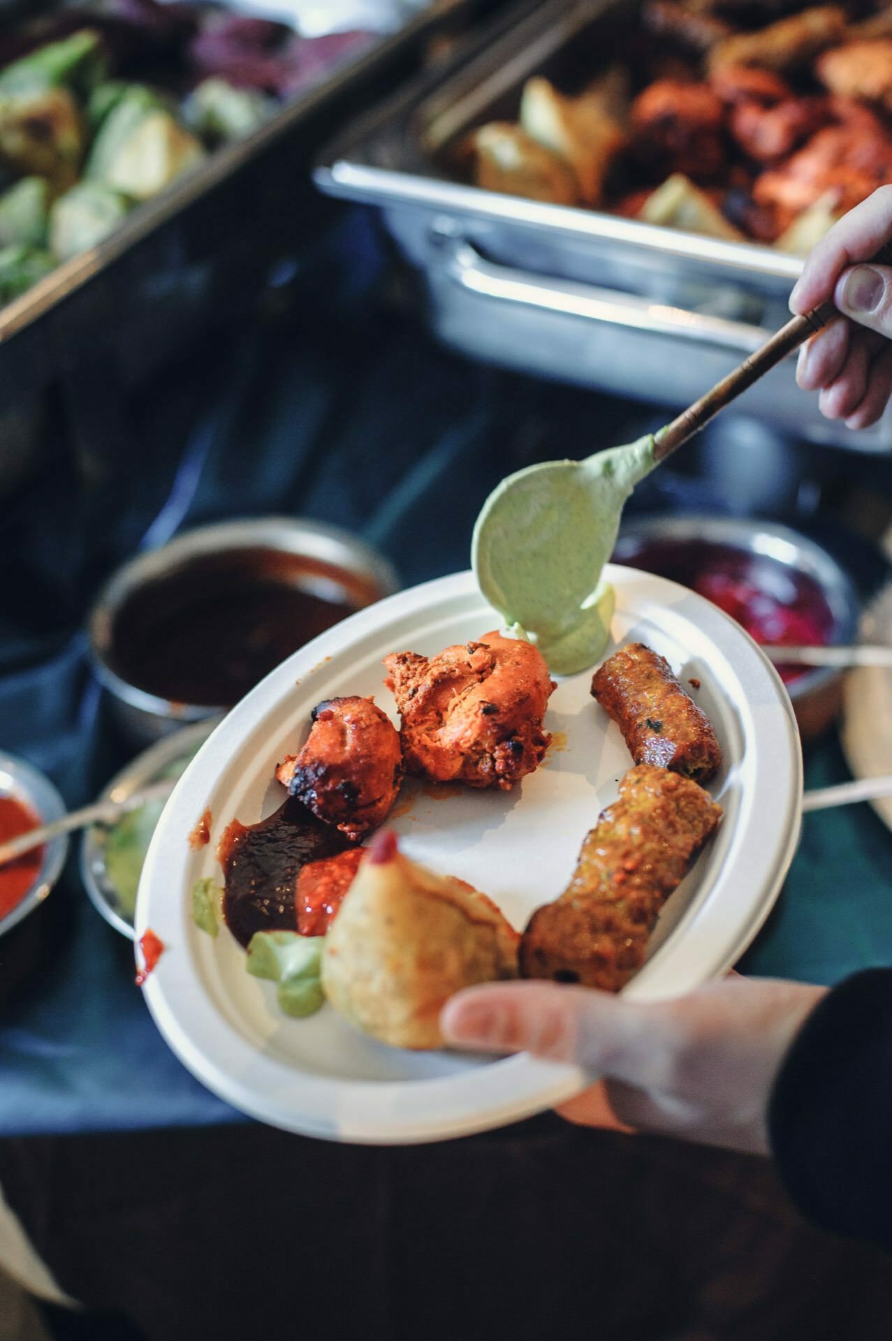 A person holding a white paper plate with various Indian appetizers, including samosa, tikka and saigon, is served green chutney with a spoon. In the background of this food fair scene, other Indian dishes and sauces are seen on trays. 