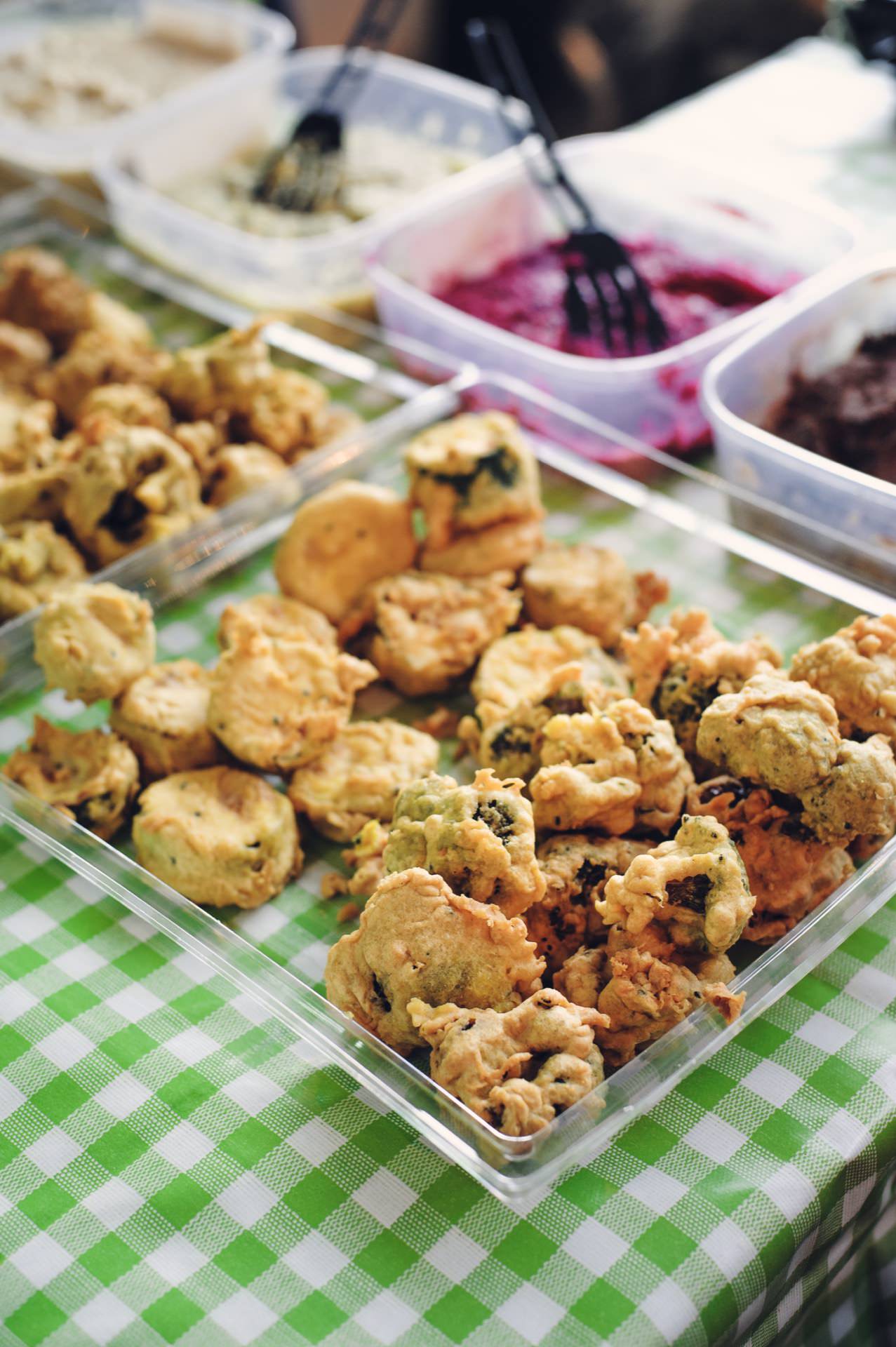 A transparent plastic tray filled with crispy, golden-fried vegetable pancakes placed on a green and white checkered tablecloth is reminiscent of a food fair. In the background you can see several plastic containers filled with various colorful dips and serving utensils. 