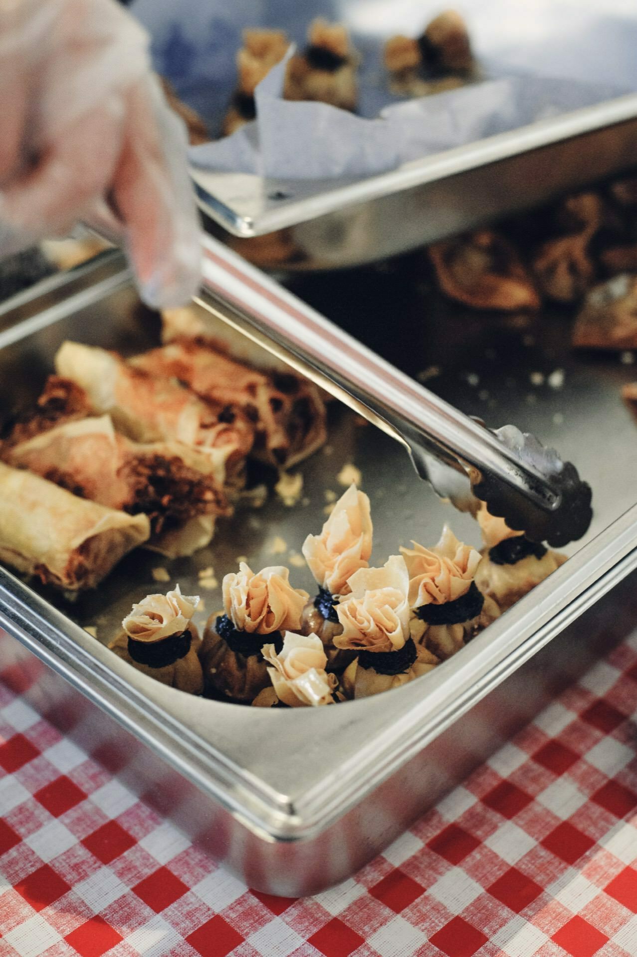 A gloved hand uses metal tongs to lift a dessert from a tray filled with a variety of pastries, set on a red and white checkered tablecloth. Additional food products can be seen in the background at the food fair, adding to the vibrant culinary display. 