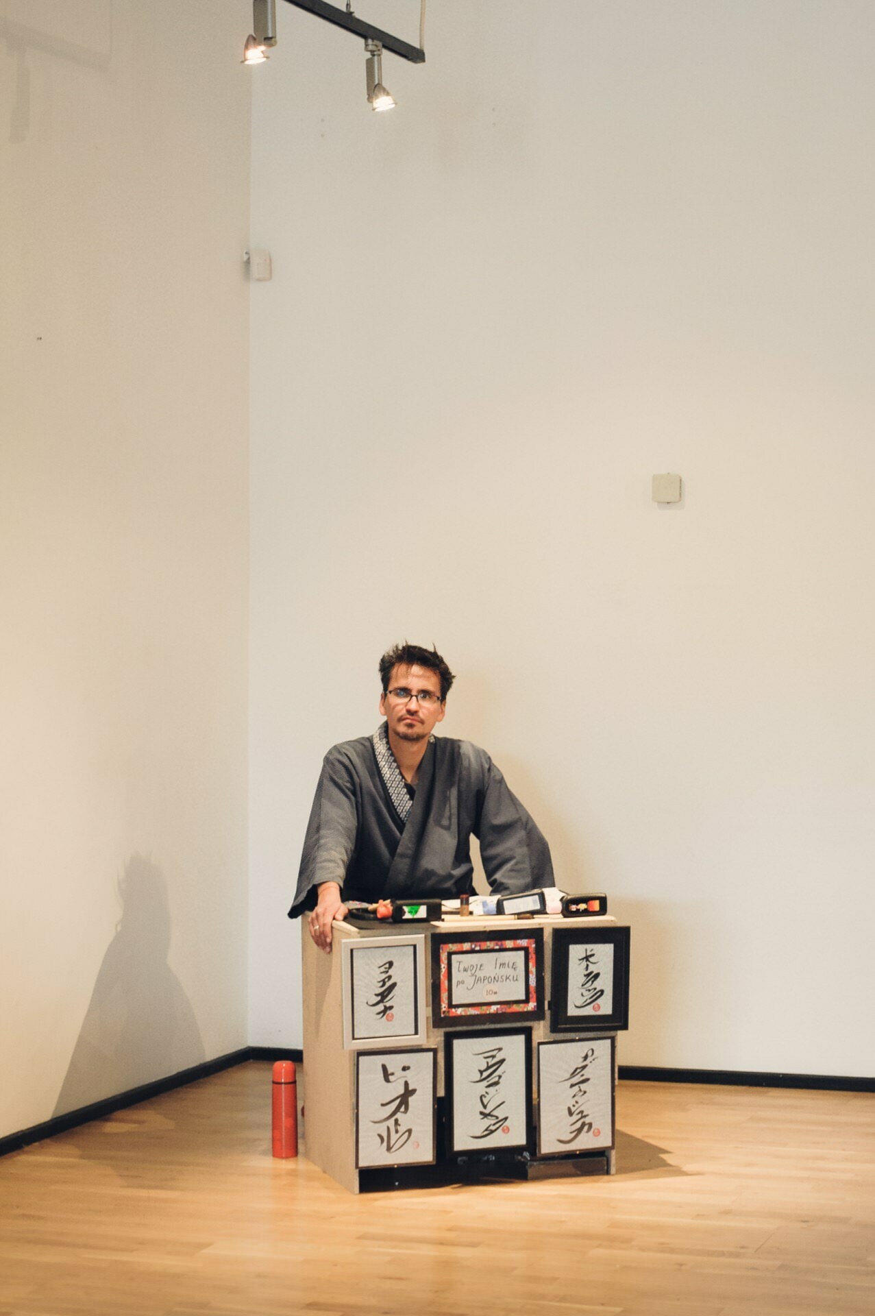 A man dressed in traditional Japanese garb stands behind an exhibition of Japanese calligraphy works on square boards. The setup is minimalist with a light wall and wooden floor, illuminated by a spotlight. In an artistic atmosphere reminiscent of a food fair, a red thermos sits on the floor to his left.  