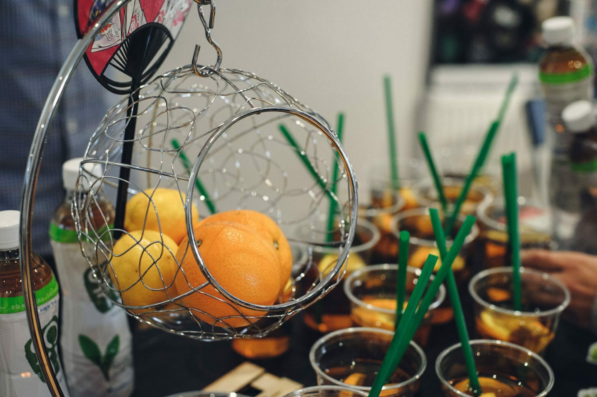 A wire basket with oranges and lemons hangs over the table. The table is set with plastic cups of iced tea and green straws. Bottled tea can also be seen on the table, among other dishes, in a sort of culinary fair, with people partially visible in the background.  