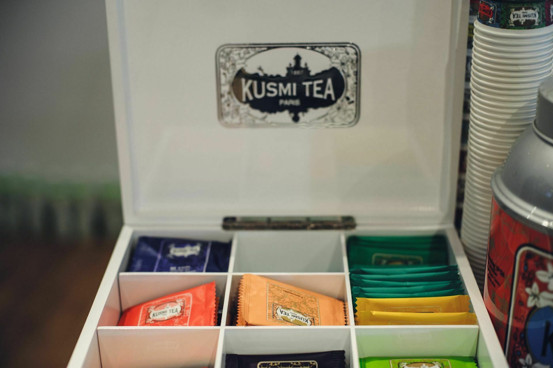 White tea box with the label "Kusmi Tea Paris" on the inside of the lid. The box contains various tea bags, each in a different color, divided into compartments. On the right side you can see stacked white cups and part of a thermos - reminiscent of displays at a food fair.  