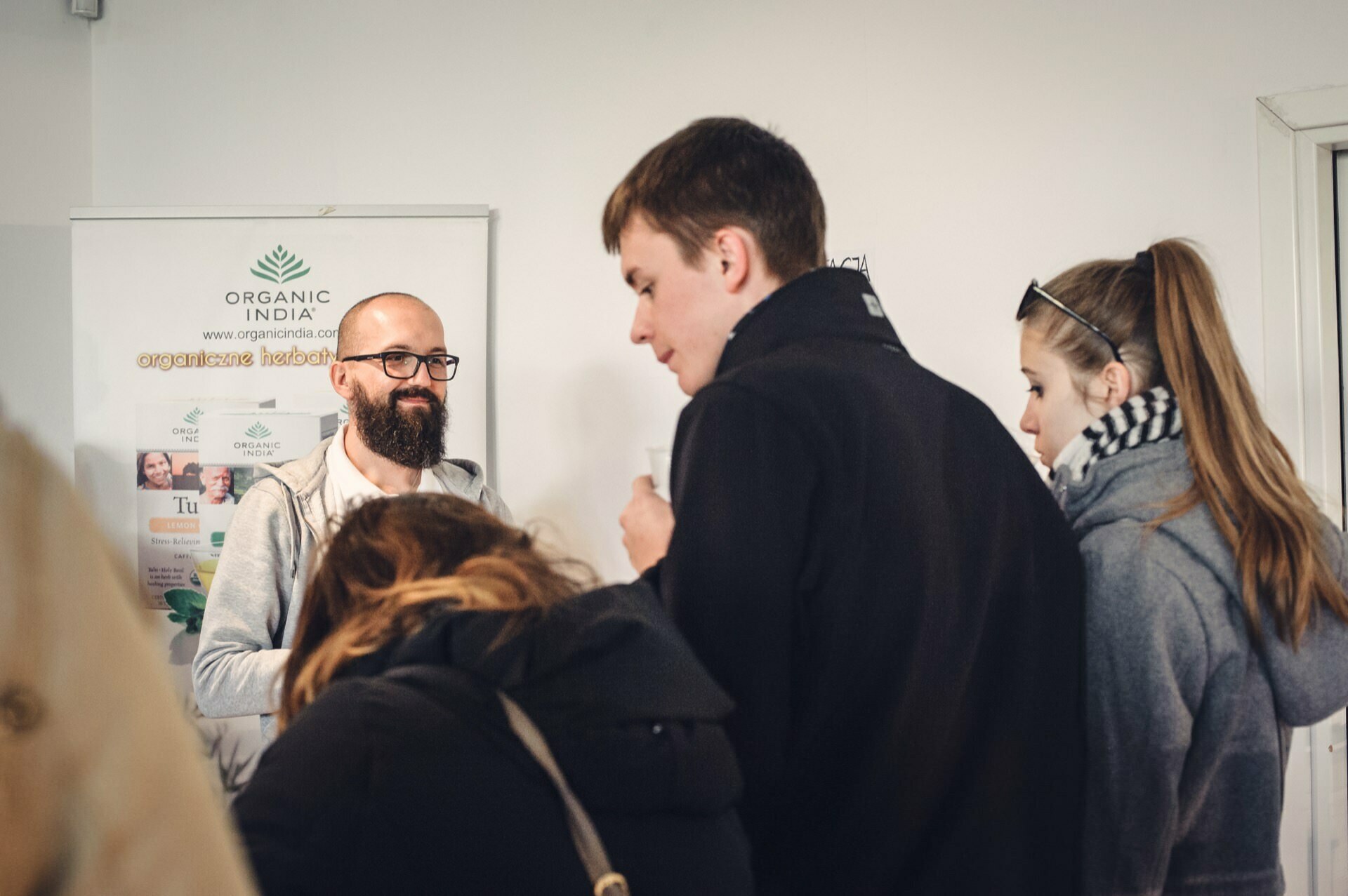 A bearded man with glasses stands next to a placard reading "Organic India" at a food fair and speaks to a small group of people. The group, made up of both men and women, are dressed in jackets and appear to be listening intently. 