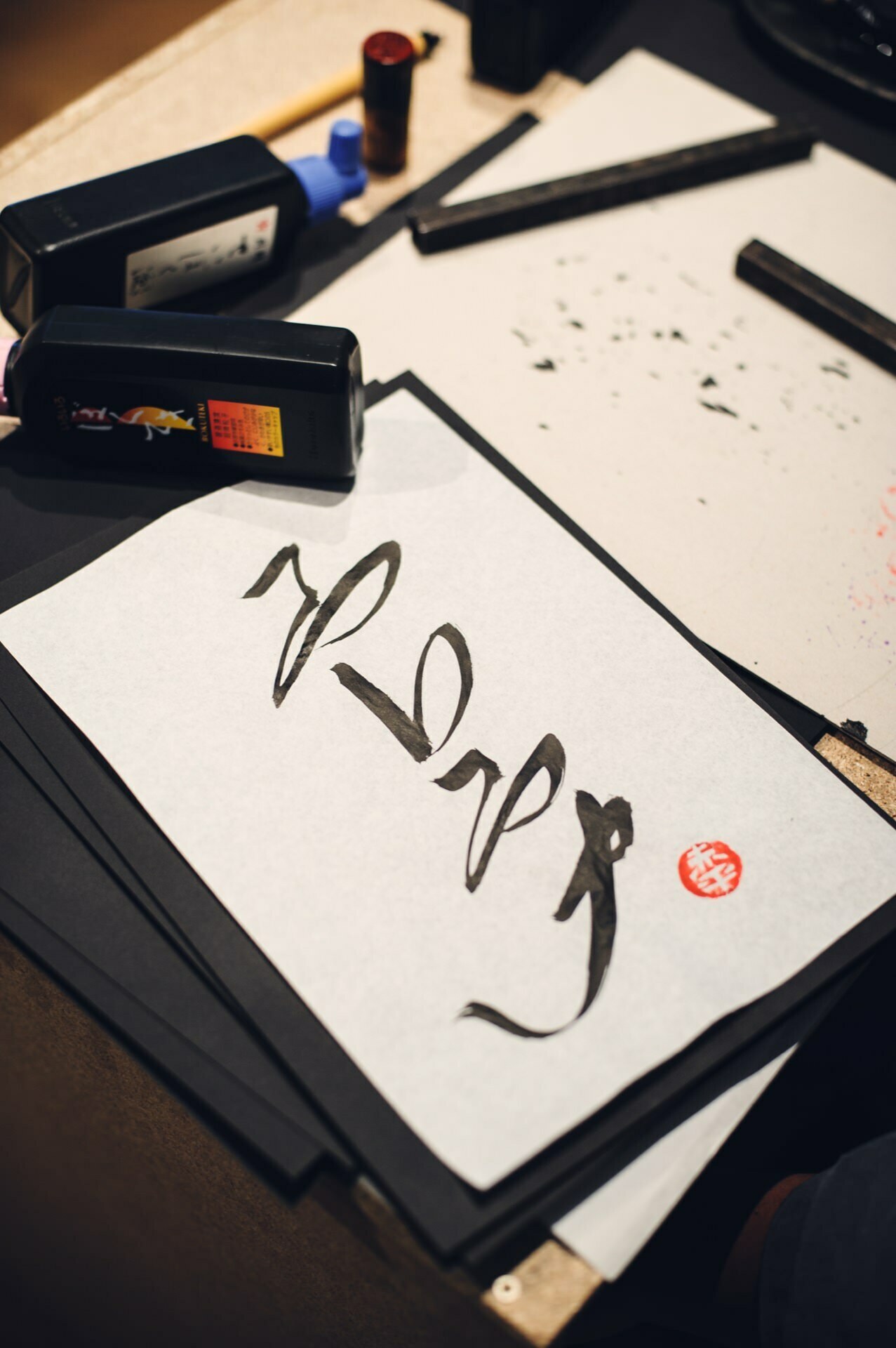 A close-up of a work area for Japanese calligraphy. On the table are several ink bottles, brushes and a sheet of paper with kanji characters made with a brush and a red stamp. The backdrop evokes the precision familiar from a cooking fair, with more sheets of paper and another brush in the ink bottle.  