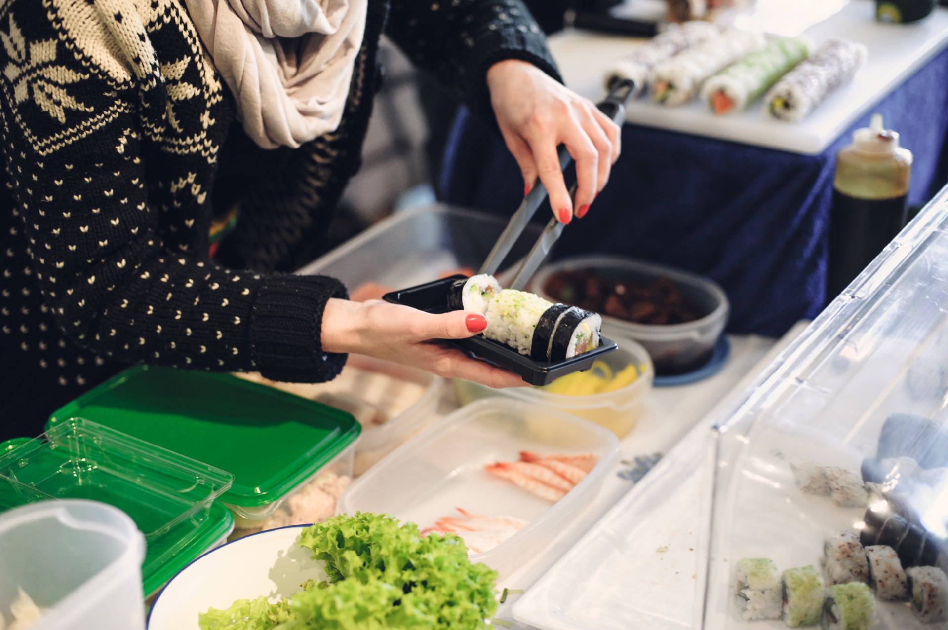 A person wearing a black sweater with white patterns and a beige headscarf cuts sushi rolls with a knife at a bustling food market. Fresh ingredients, such as lettuce and shrimp, are seen in open containers on the table. More sushi rolls and equipment can be seen in the background.  