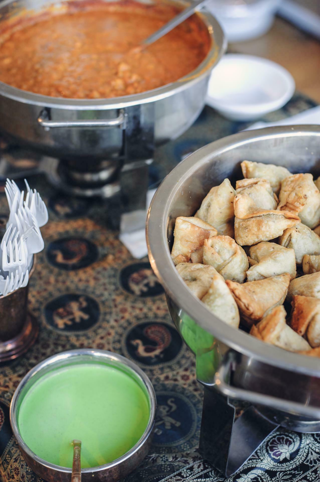 The Indian cuisine display at the food fair includes a metal container with samosas, a bowl of chickpea curry, a small bowl of green chutney and a cup with plastic forks, all arranged on a patterned tablecloth.