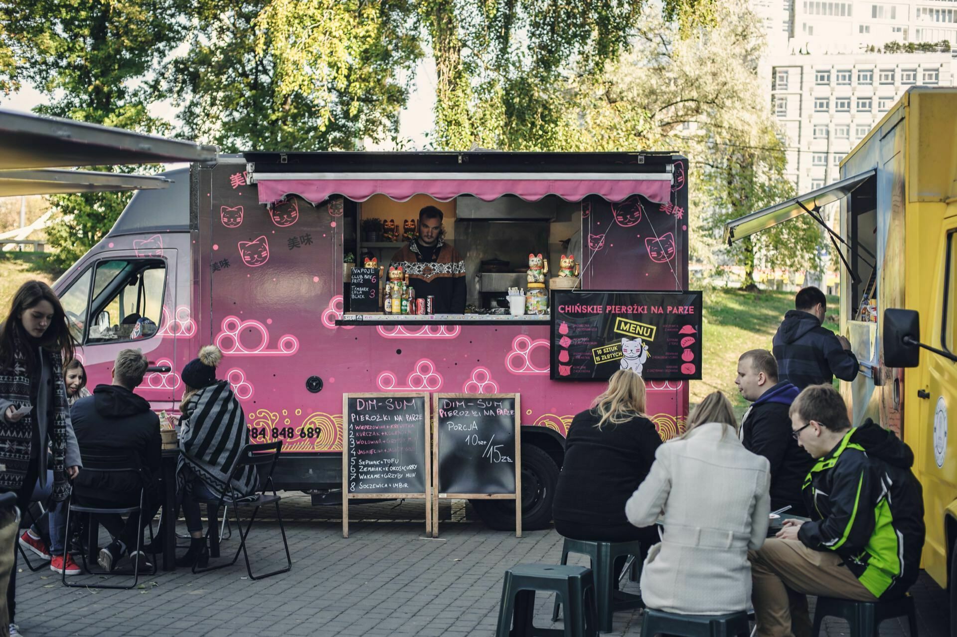 Photo of a food truck with a pink body and a cat motif parked on the pavement at a food fair. People are sitting on stools and benches in front, eating or placing an order. On a sunny day, trees and a multi-story building can be seen in the background.  