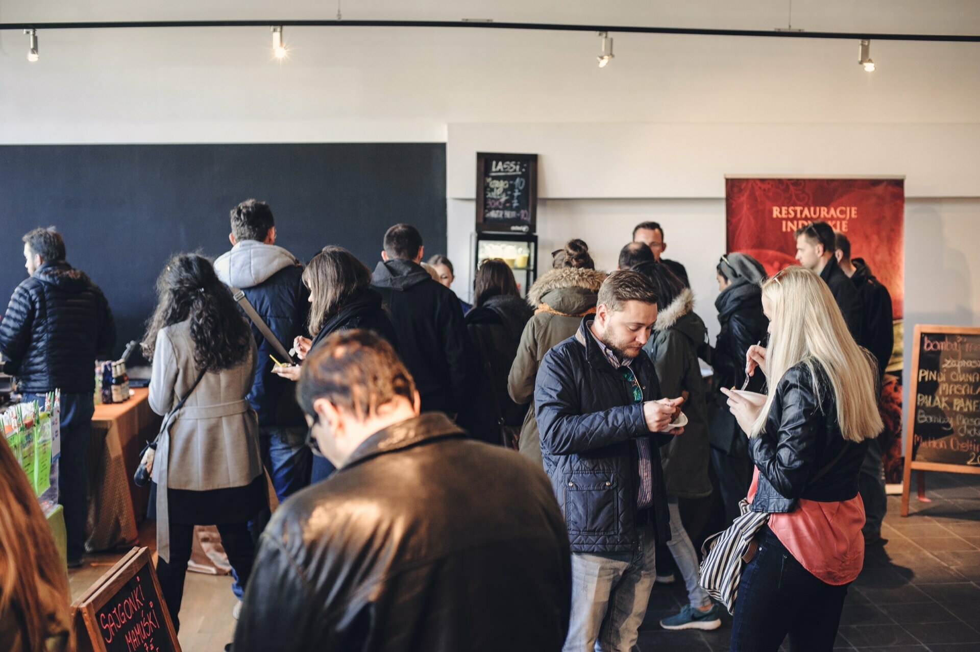 A group of people gather in what appears to be a casual room, reminiscent of a food fair. Some are sitting, others are standing, and many are using their phones. A red sign in the background reads "Restaurants." There are tables with various items posted on them.   