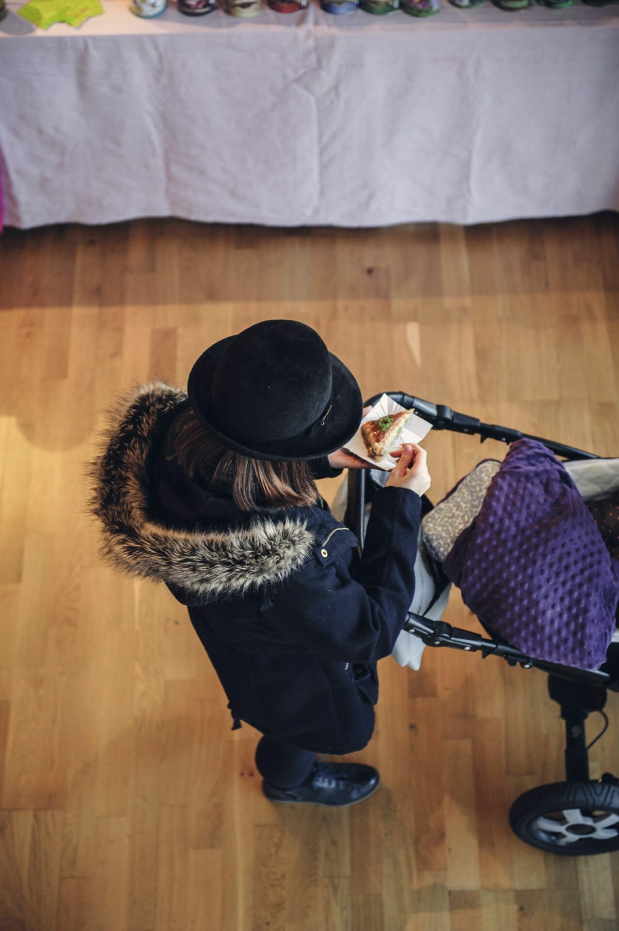 From above, a person wearing a black hat and a fur-trimmed hooded coat can be seen holding a piece of food. They are standing next to a cart with a purple blanket on the wooden floor of the food fair, in front of a table covered with a white tablecloth. 