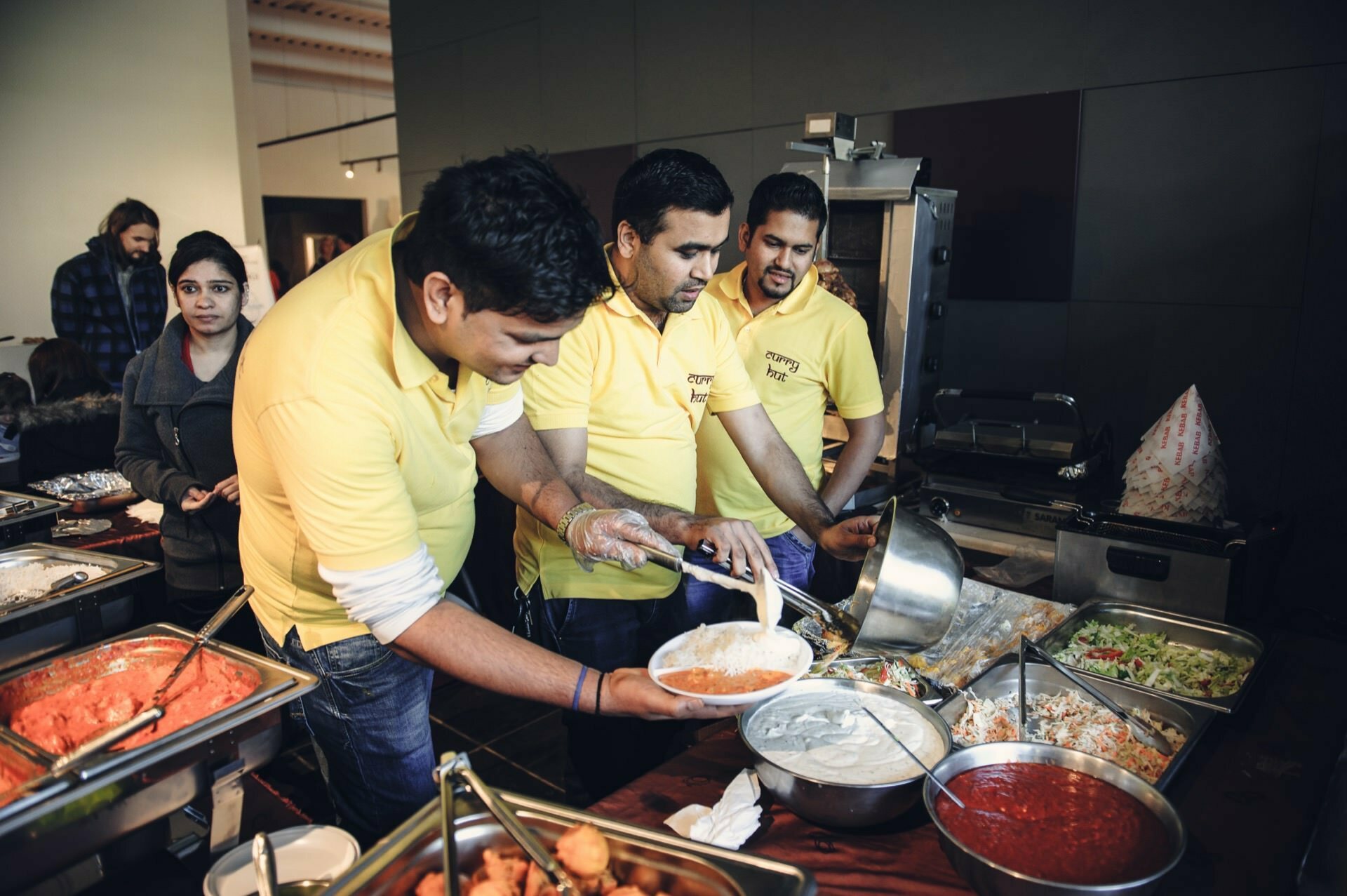 Three people in yellow shirts serve food from a buffet during an event held indoors. They put various dishes, including curries and salads, on plates. Other people and buffet trays are visible in the background. The atmosphere at the food fair is lively and engaging.   