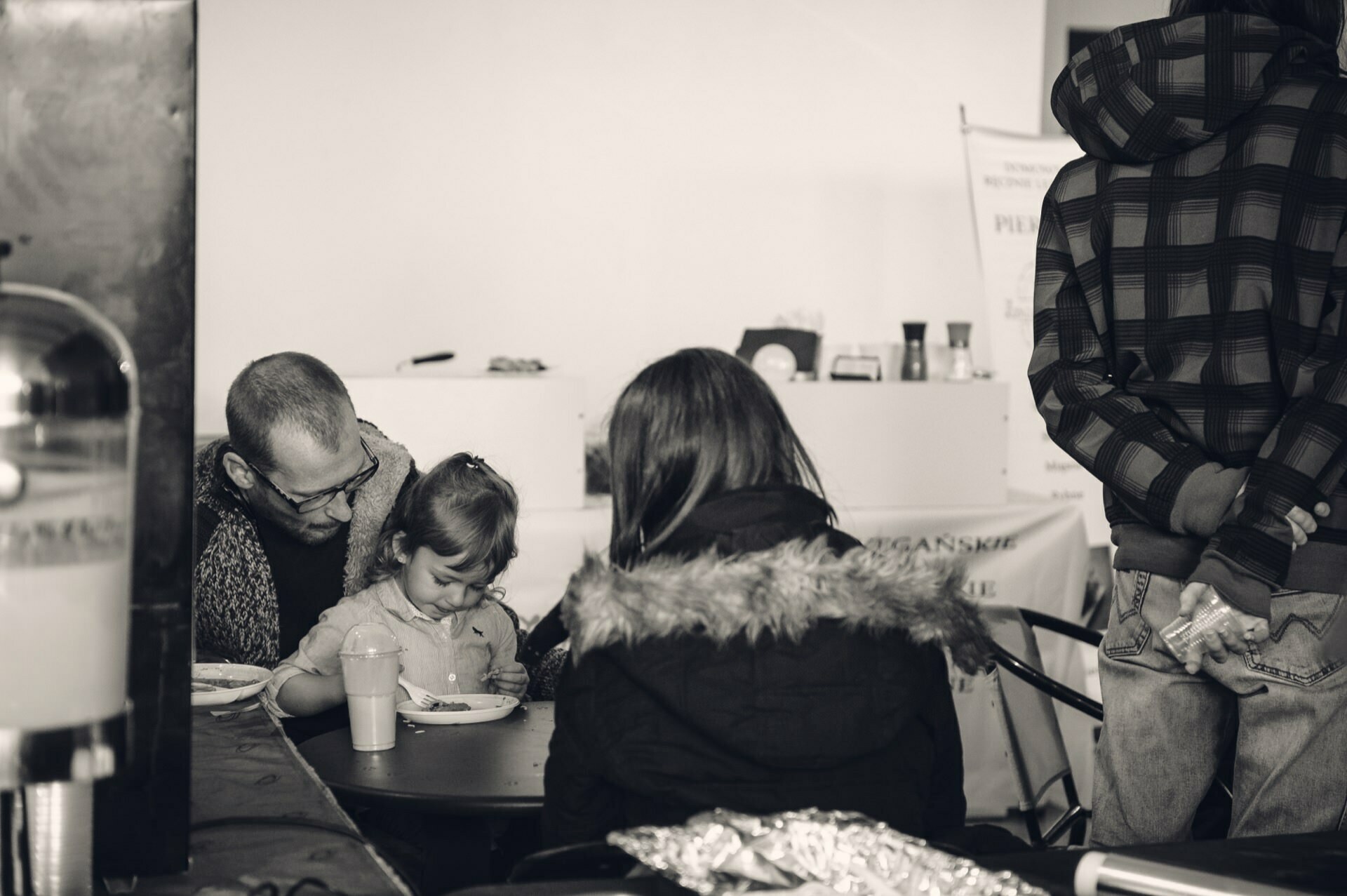 The grayscale photo shows a family at a table, probably at a food fair. On the left sits a man wearing glasses and a beard, helping a small child with food. A woman in a fur-trimmed coat with a hood sits in the middle, with her back to the camera. To the right stands another person in a plaid hooded sweatshirt.   