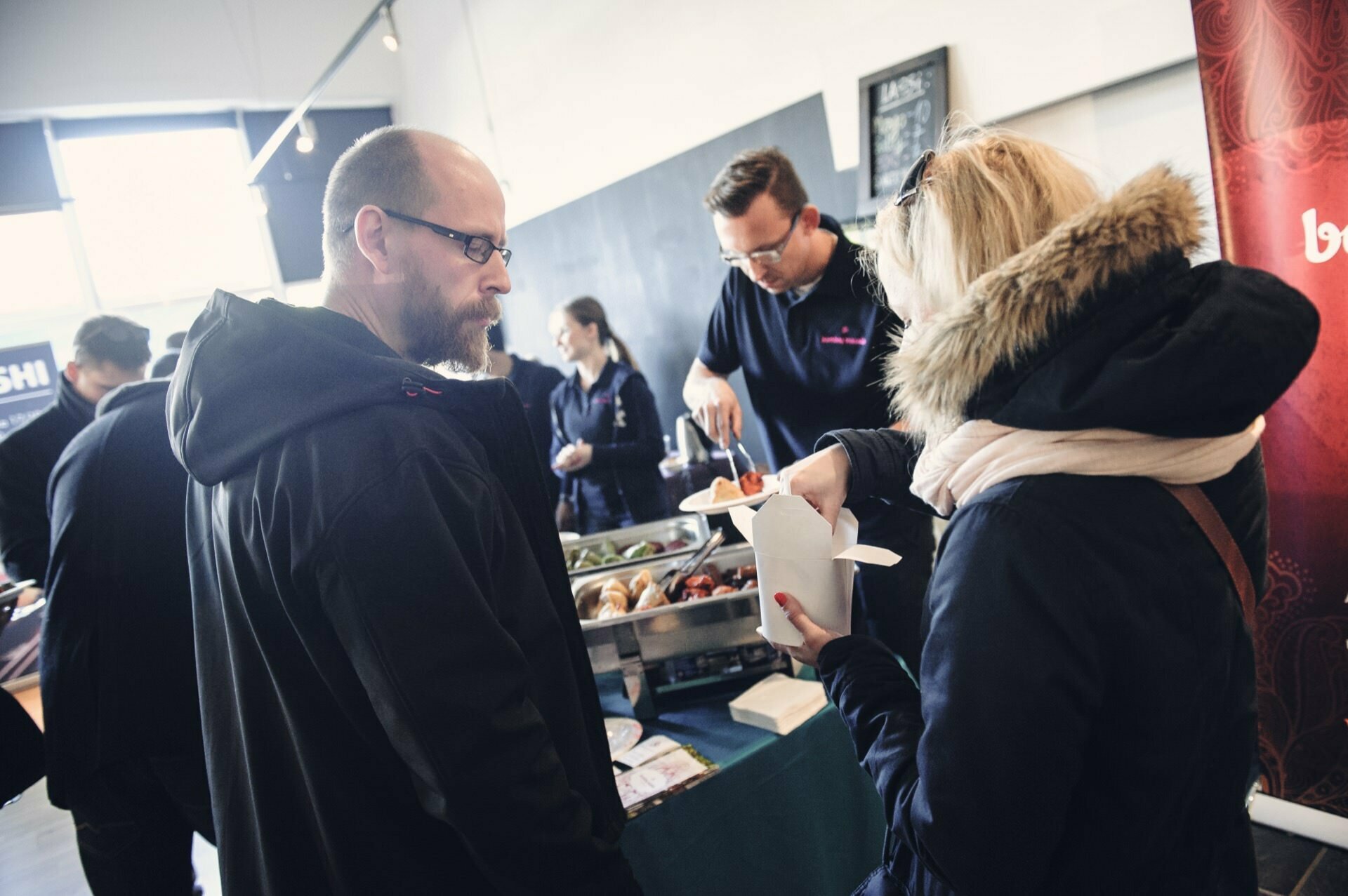 A man at a food stand serves food to two people wearing winter jackets. The woman is holding a take-out box while the bearded man looks on. Behind them, at various food fair booths, stand others. The atmosphere is casual and busy.   