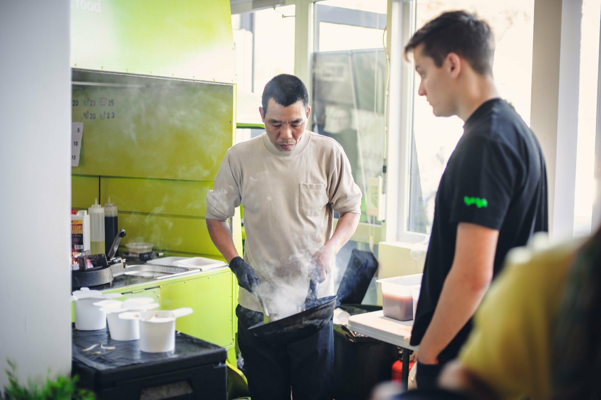 A gloved cook prepares food at the stove, and steam rises from a pan in the small green-tiled kitchen. A young man in a black shirt stands nearby and watches intently, resembling impatient culinary fair attendees. Bright natural light streams in through the windows.  