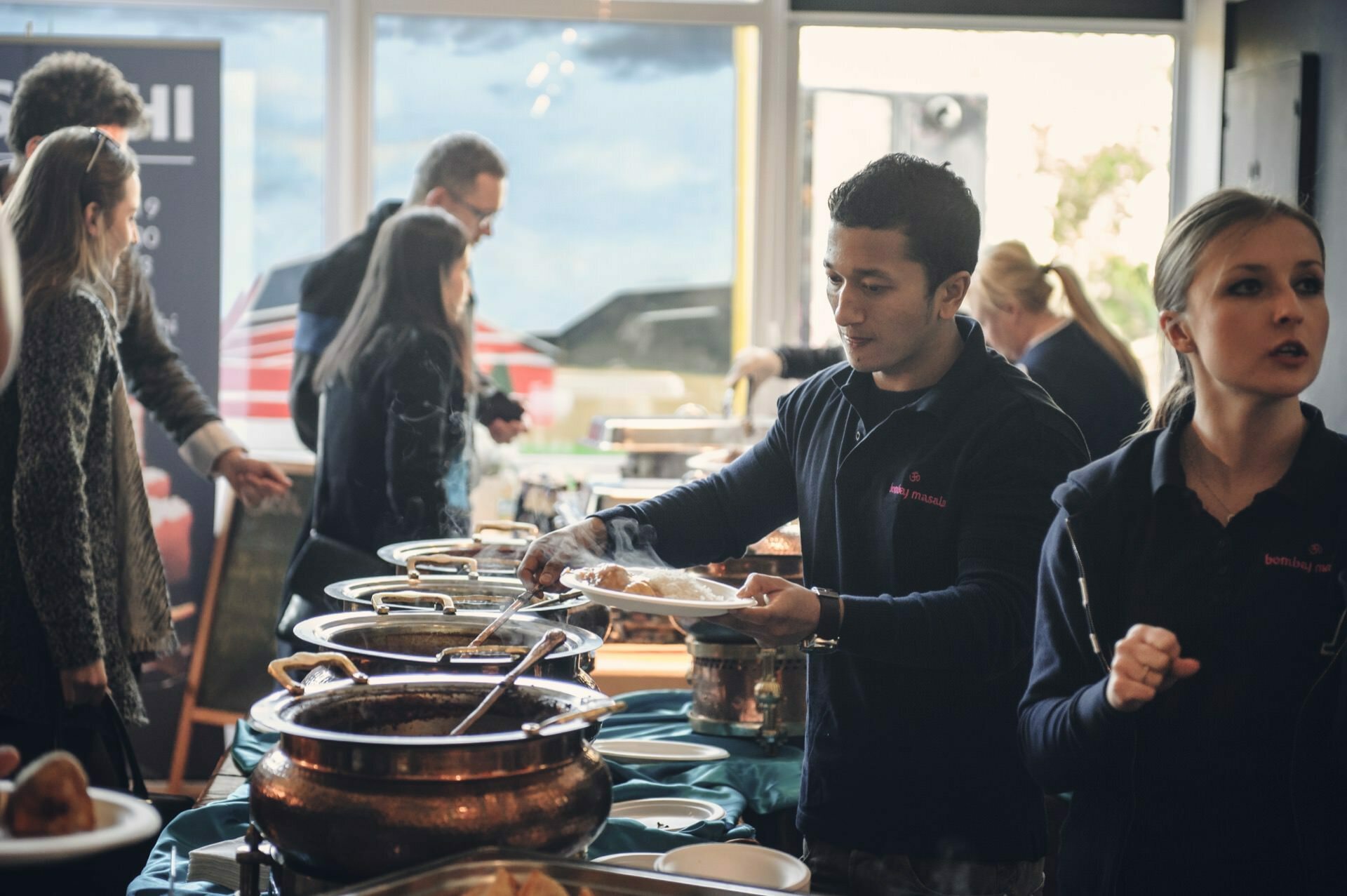 At a food fair, a group of people are serving themselves food from a buffet table filled with a variety of dishes in food detractors. Two people in the foreground serve food on plates while more people can be seen through a large window in the background. 