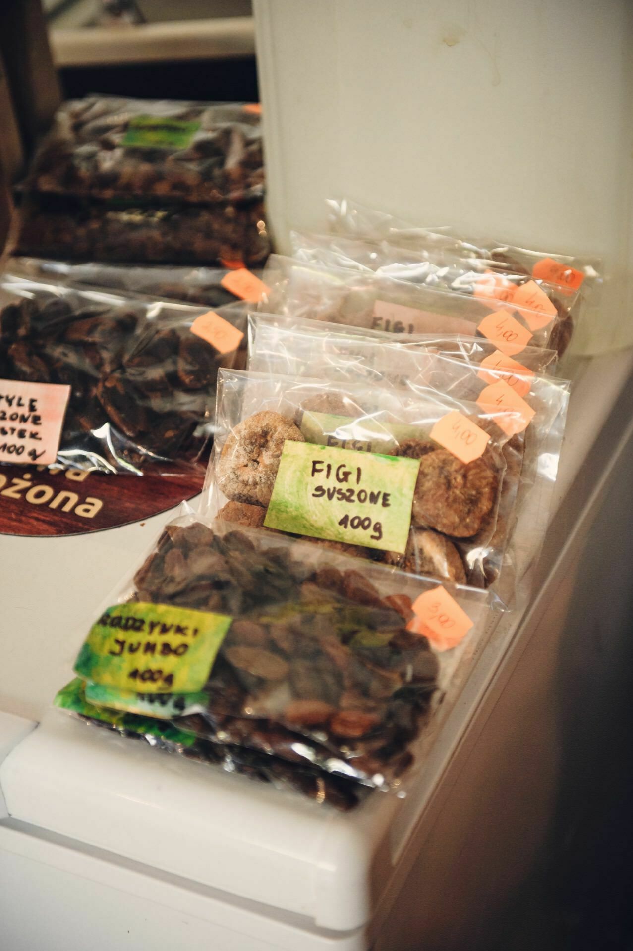 At food markets, various packages of dried fruits are displayed on the counter with signs in Polish. For example, there are dried figs and other dried fruits in plastic bags, and orange stickers with prices on each package. 