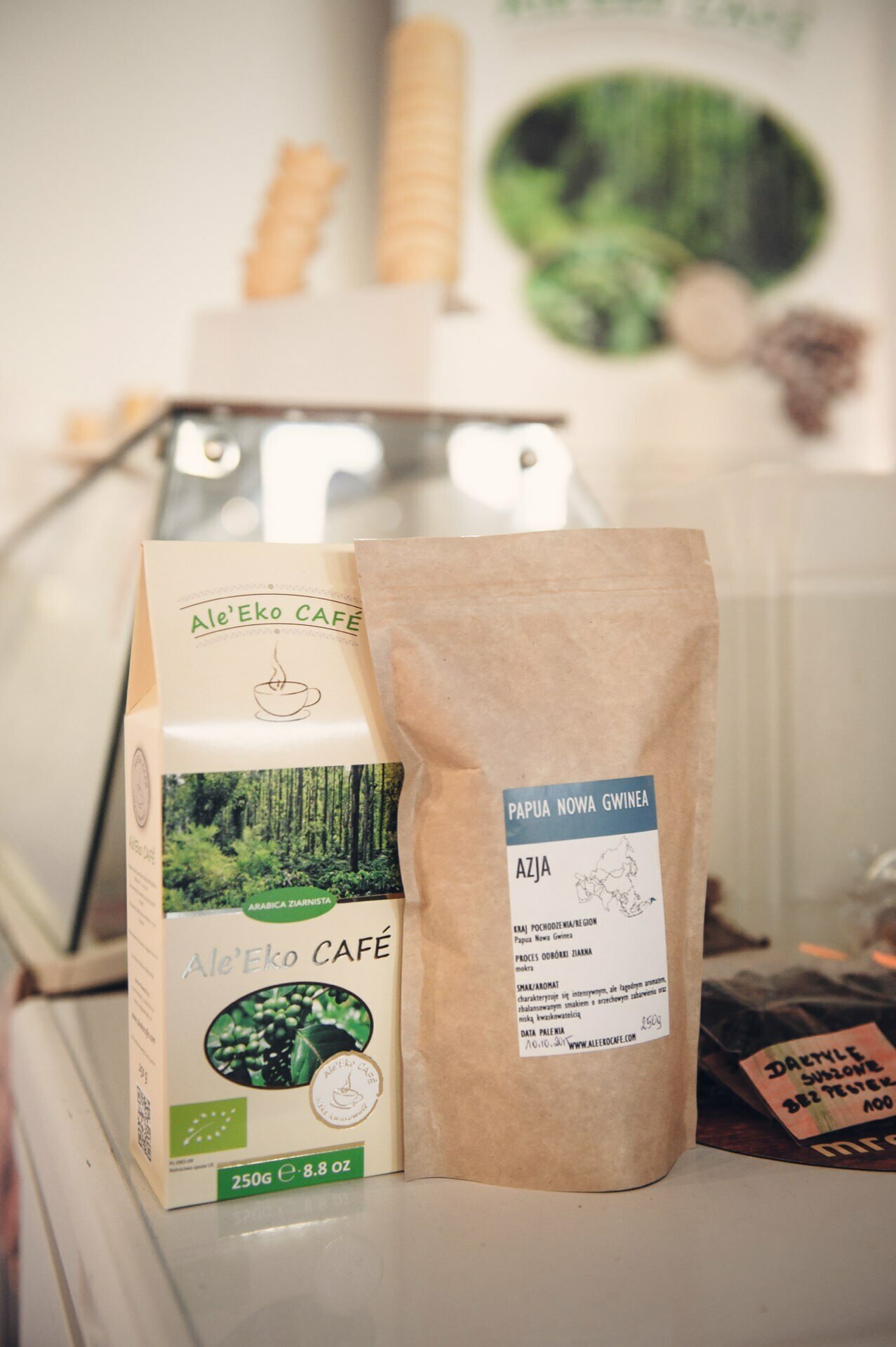 Two packages of coffee are on display at the culinary fair: one in a green and white box labeled "Ale'Eko Café" and the other in a brown paper bag labeled "Papua New Guinea Asia." Both packages are on a white surface with a fuzzy background depicting the café's decor. 