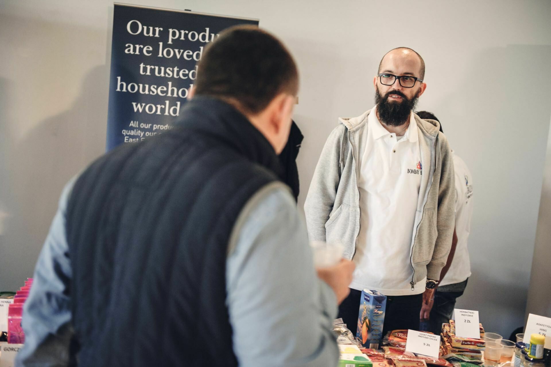 At a food fair, a man in a white polo shirt stands behind a table displaying various products and talks to another person, facing away from the camera. A placard in the background proclaims: "Our products are recognized and trusted by households.... 