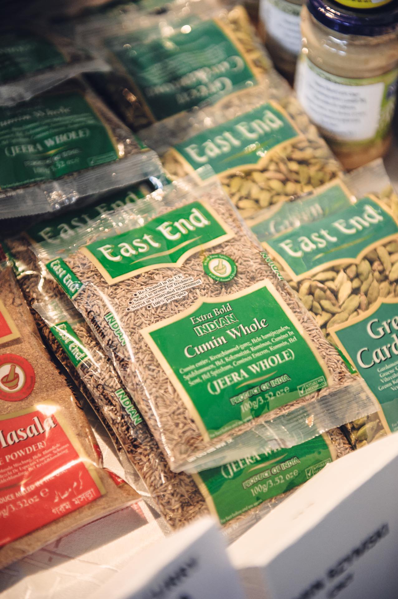 A close-up of various packaged spices of the East End brand displayed on a shelf at a culinary fair. Visible among the packages are whole cumin, green cardamom and garam masala powder. A jar containing a mustard-colored substance can also be seen in the background.  