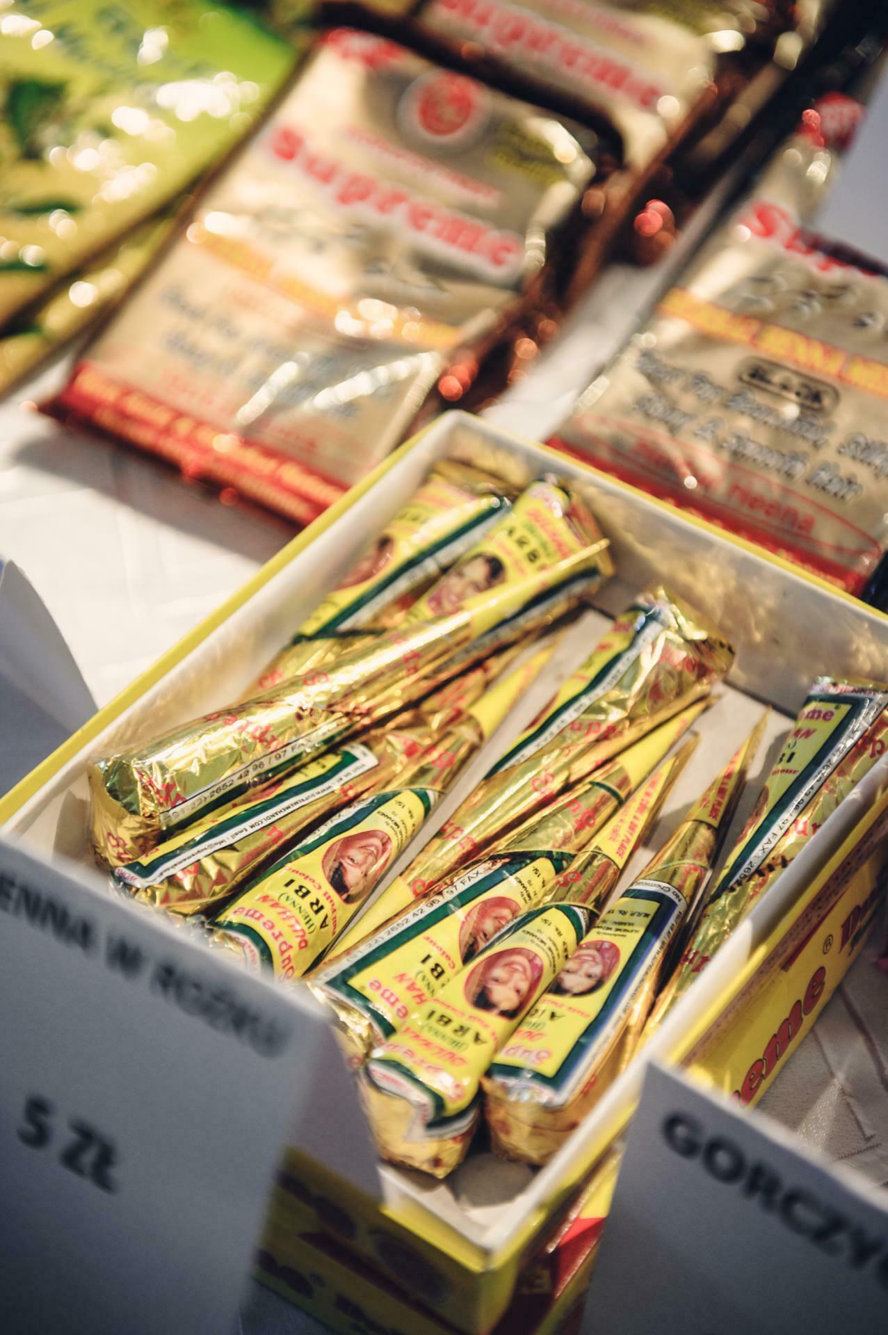 A box of tubes in yellow and gold packaging, displayed on a table at a culinary fair. The tubes are labeled with images of faces and text. The background includes additional packs in gold and red, which also feature faces. In the foreground, a label with the price of "5 zloty" is visible.   