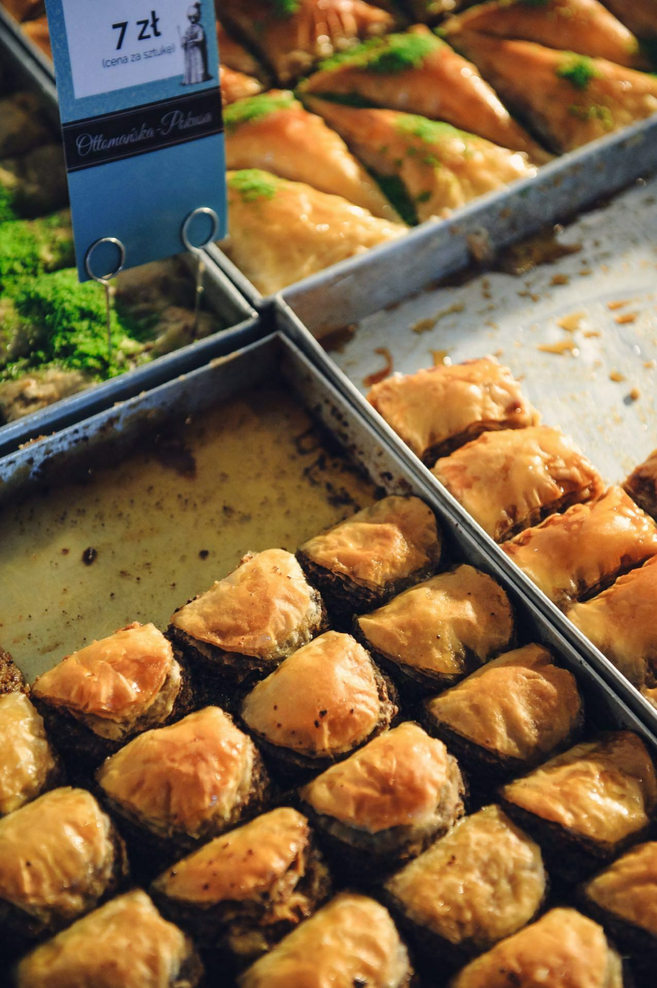 A display of various baklava baked goods on a tray at a bakery during a culinary fair. The golden brown squares are neatly arranged, some topped with green pistachios. An inscription indicating the price of 7 zloty per piece is visible in the upper left corner.  