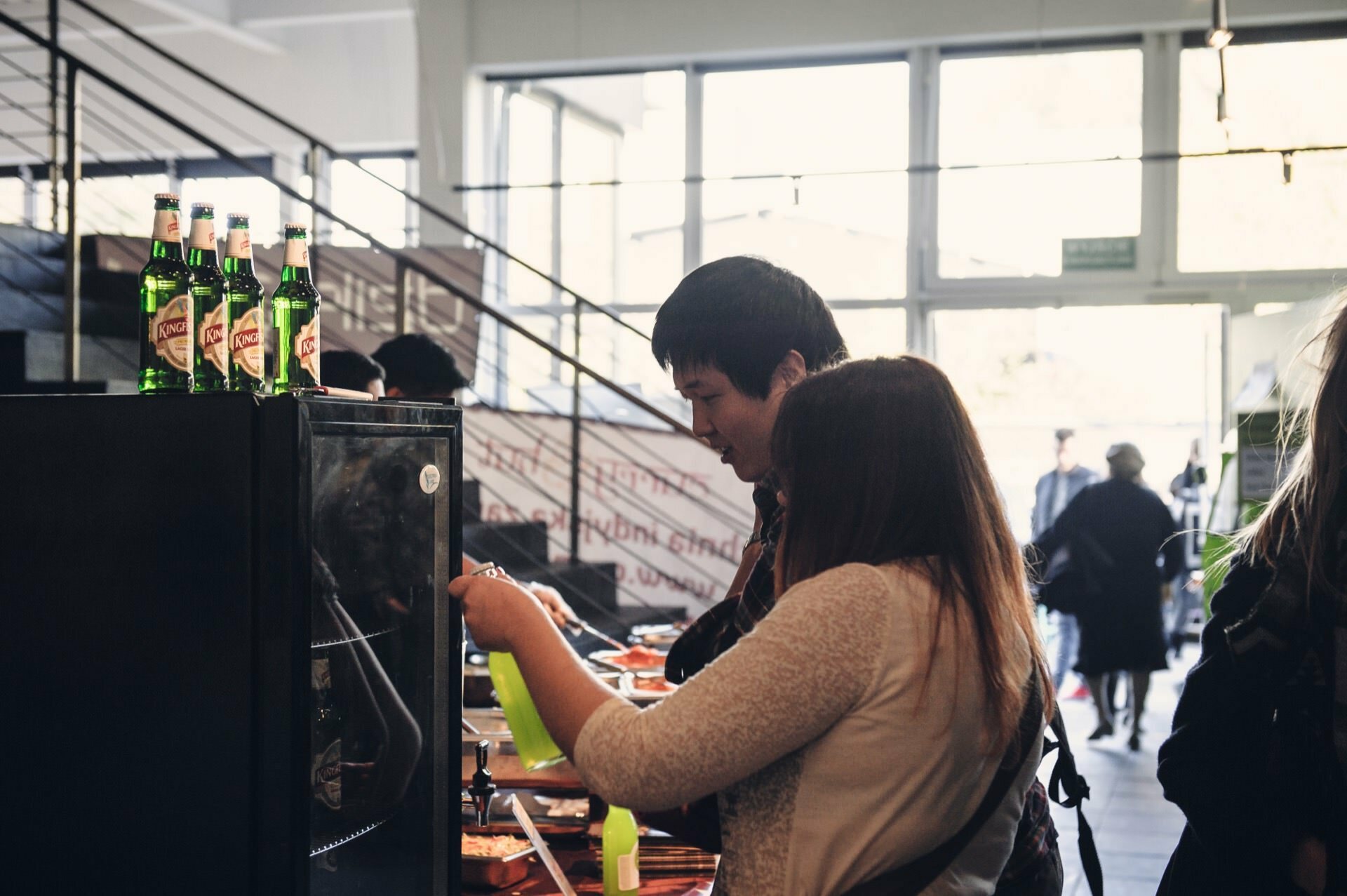 Two people, a man and a woman, serve themselves food from a table in a brightly lit café or dining room, reminiscent of a food fair. Bottles of drinks can be seen on a nearby refrigerator. In the background, near the entrance, other people can be seen.  