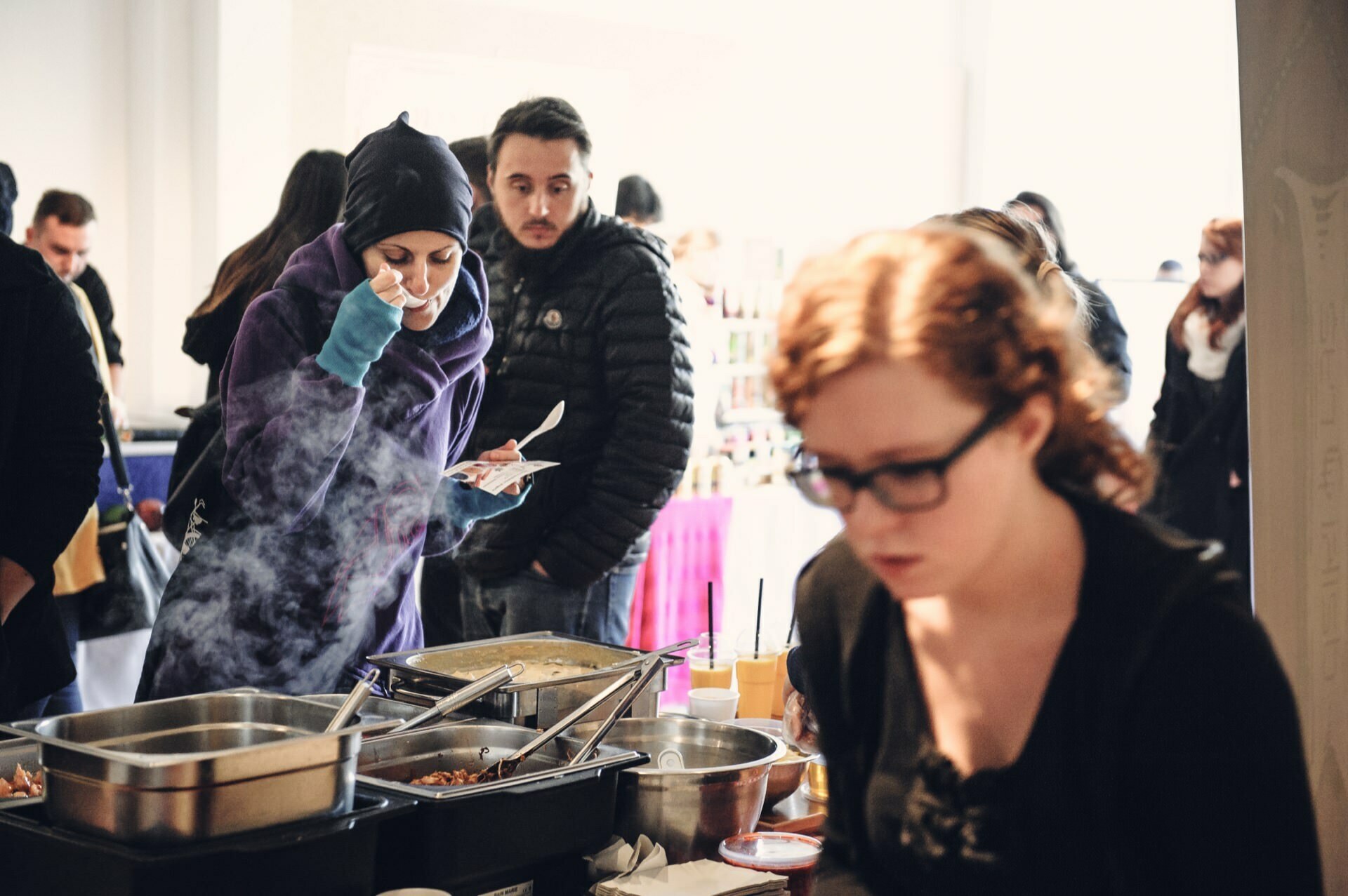 People gather around a buffet table at an indoor food market. A woman in a headscarf tries the food, others wait in line. In the foreground, a woman with glasses is focused on serving, while steam rises from the dishes on the table. The atmosphere is casual and busy.   