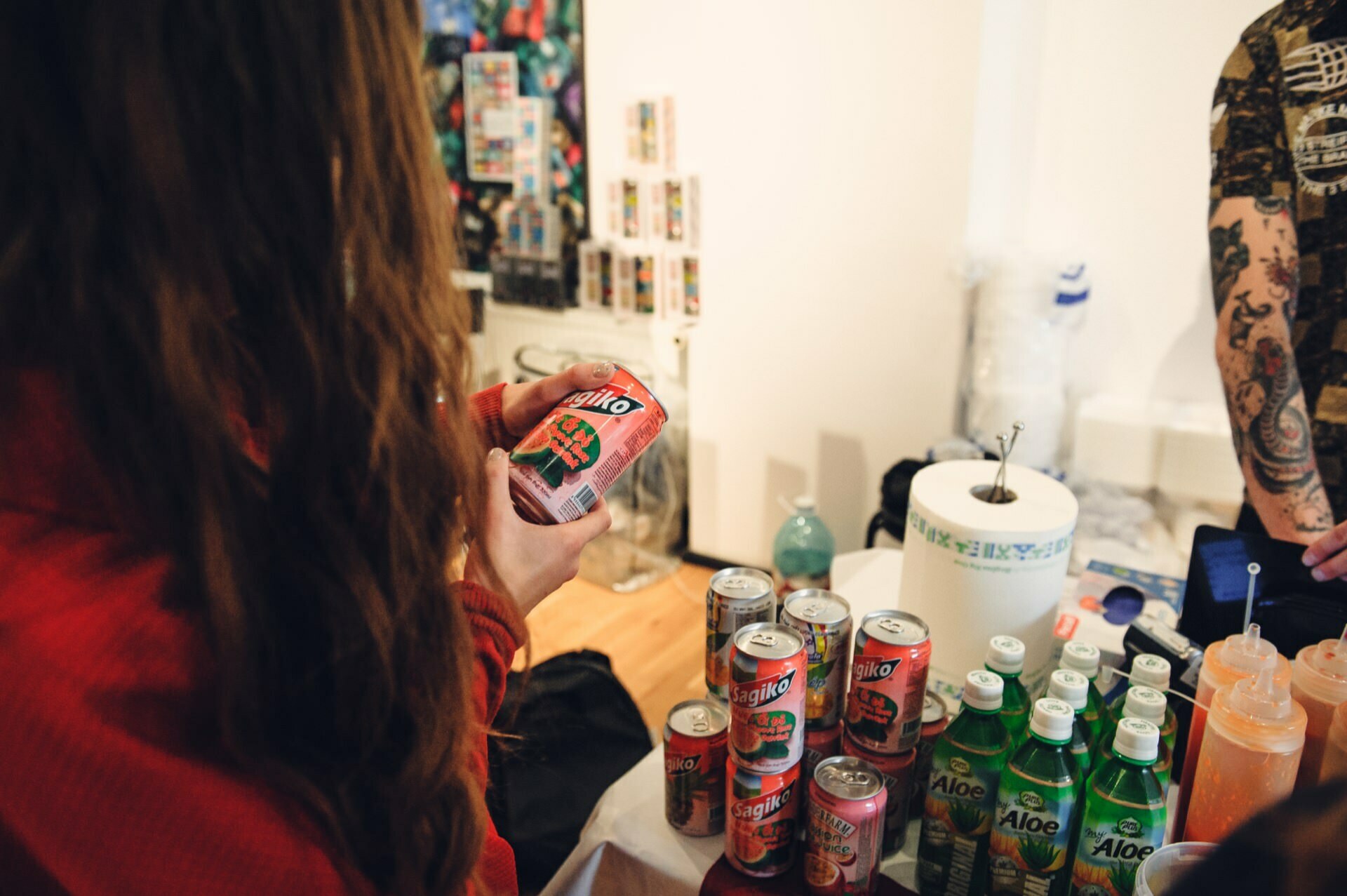 At the food fair, a person in a red shirt is holding a can of soda, and several cans of sodas and aloe vera drinks are displayed on a table. Various objects and their tattooed arm are visible in the background. 