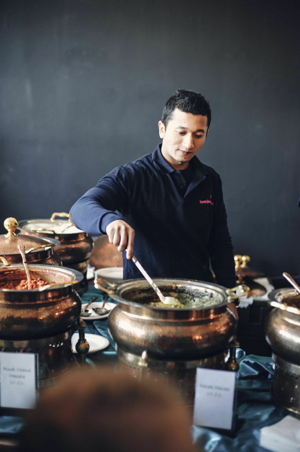 A person serves food from a large pot at a buffet table. There are several similar pots on the table, each containing different dishes. The person is wearing a dark long-sleeved shirt and appears to be at a food fair or other catering or restaurant event.  