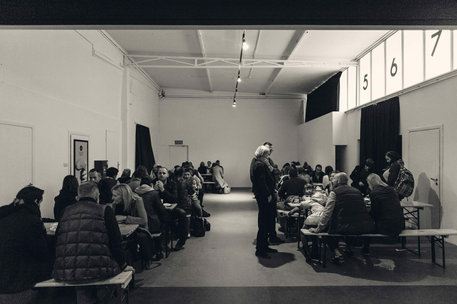 The monochromatic image shows a large room filled with people sitting on benches along long tables, reminiscent of a food fair. They appear to be engaged in conversation or small group activities. The room has high ceilings with exposed lighting and large numbered windows along one wall.  