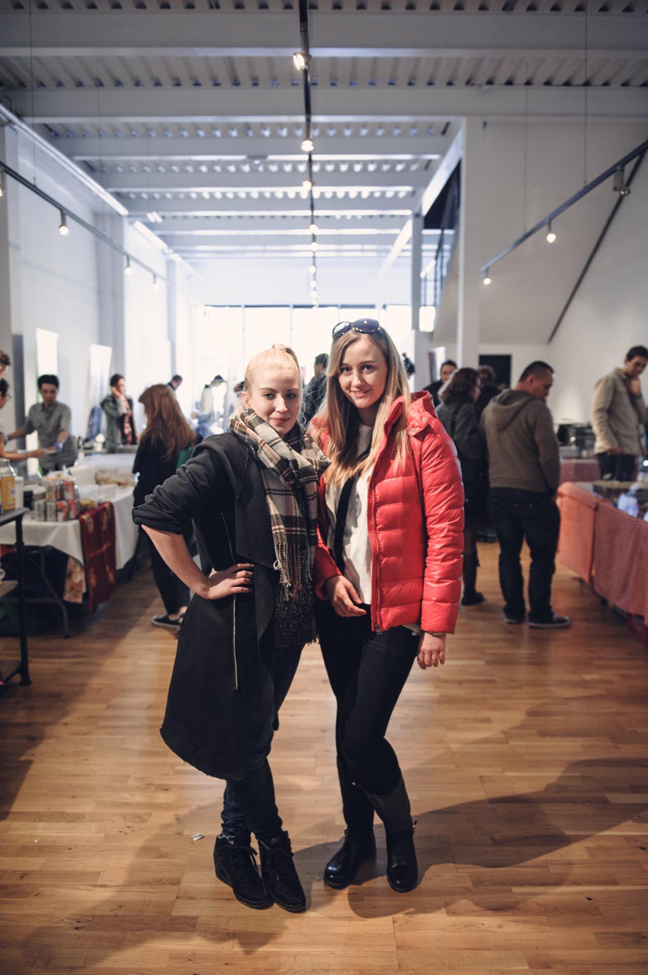 Two women pose and smile in a well-lit marketplace. One is wearing a black outfit with a plaid scarf and the other a red jacket. Stalls and people can be seen in the background. Wooden floors and a high, exposed ceiling add to the charming atmosphere of the food market.   