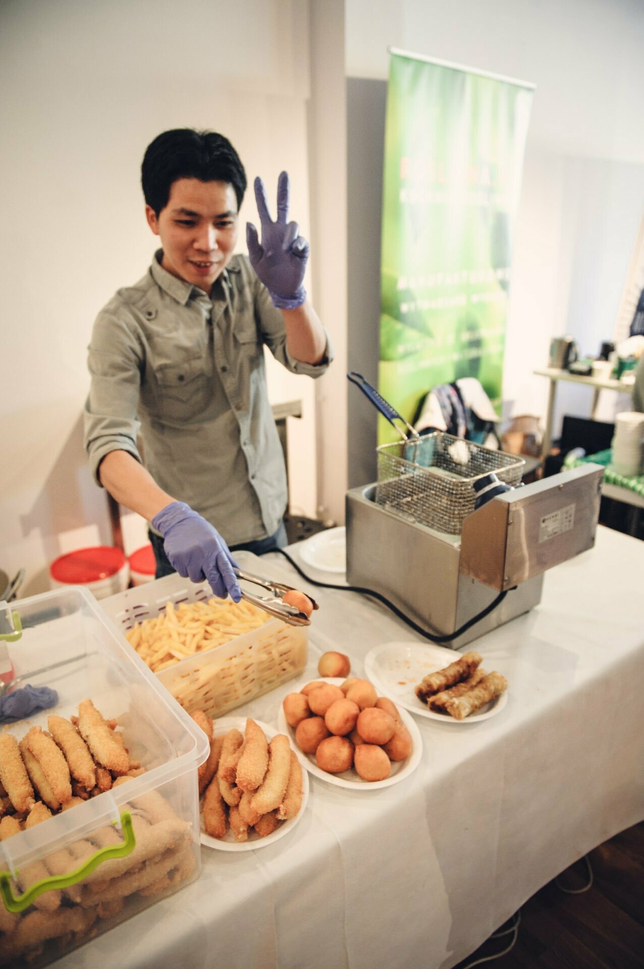 A gloved person is standing behind a table at a food fair displaying various fried foods, including French fries, dough balls and breaded dishes. They hold tongs in one hand and gesture with the other. Next to them on the table is a commercial fryer.   