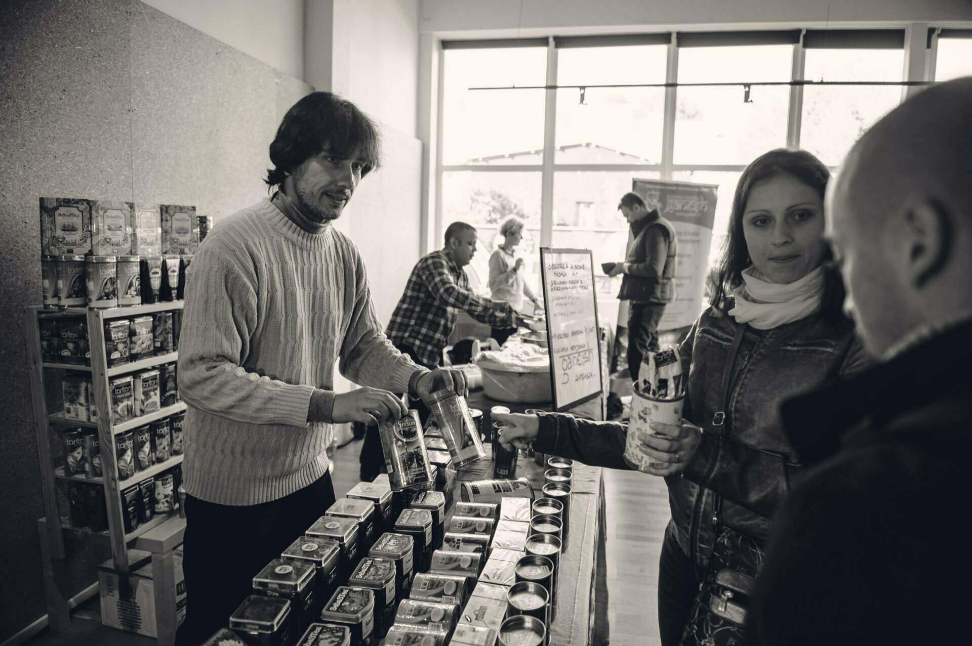 Black and white image of a market scene in a room. A sweater-clad vendor serves a product to a customer across a table filled with various packaged goods. At the bustling food market, other shoppers and vendors are visible in the background. The atmosphere seems casual and busy.   