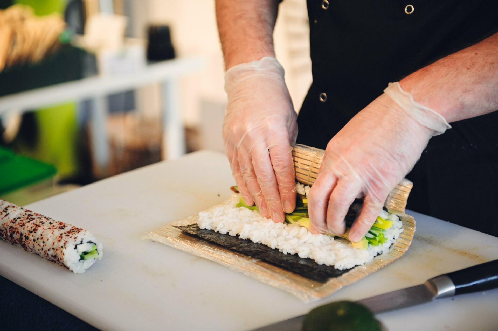 A person wearing gloves prepares sushi rolls at a food fair. They use a bamboo sushi rolling mat to roll seaweed, rice and vegetables together. The finished sushi roll and knife lie next to them on a white cutting board.  