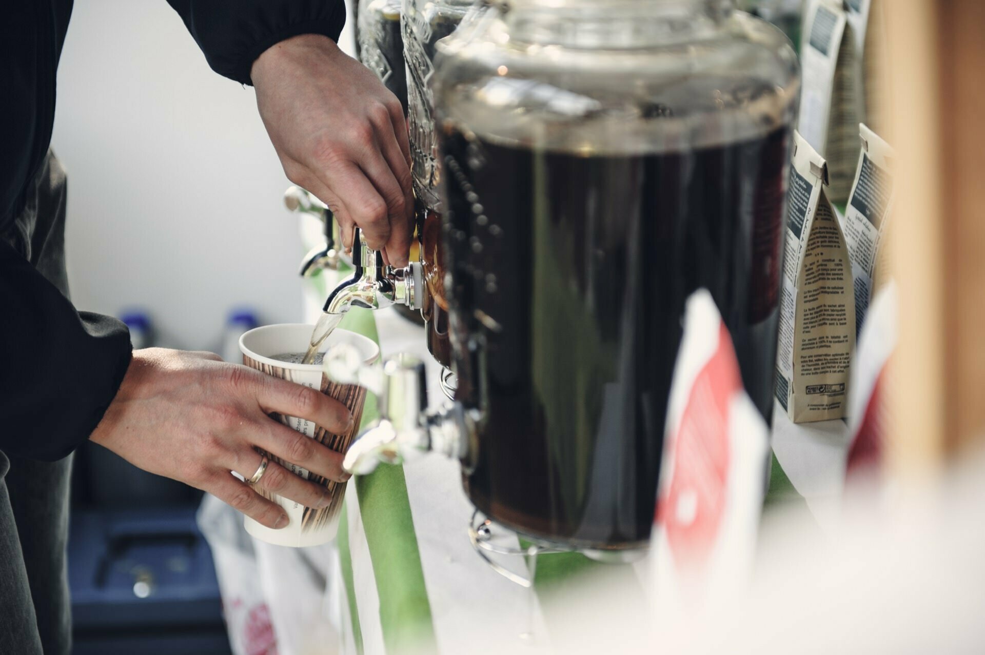 Hands can be seen filling a disposable paper cup with a dark liquid, probably coffee or tea, from a large transparent dispenser with a faucet at a self-service stand at a food fair. Various packages and containers are visible in the background. 