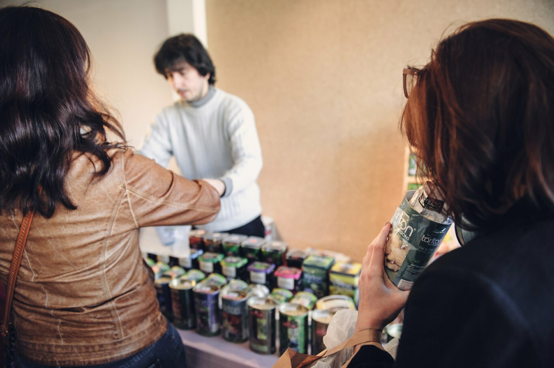 People are shopping at a stand with cans of various products displayed on a table. A salesman in a white sweater helps a customer while another person in a brown jacket picks up a can. At a food fair, a person wearing glasses carefully watches the scene while holding a can.  