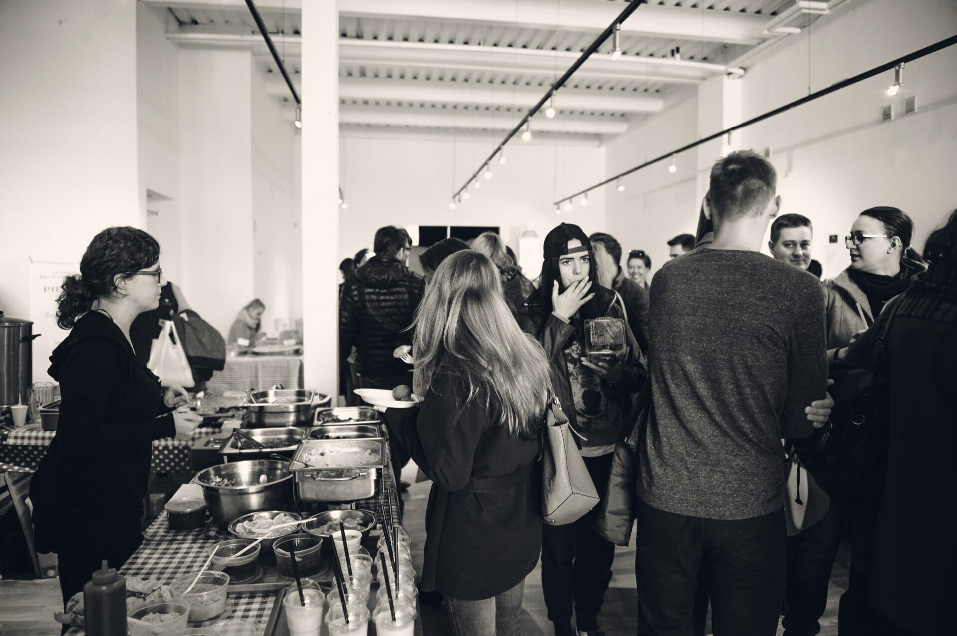 A crowded indoor gathering where people stand around tables laden with various foods, like a bustling culinary fair. Some people serve or select food, while others talk and eat. The atmosphere is casual, with a buffet, creating a lively and communal atmosphere.  