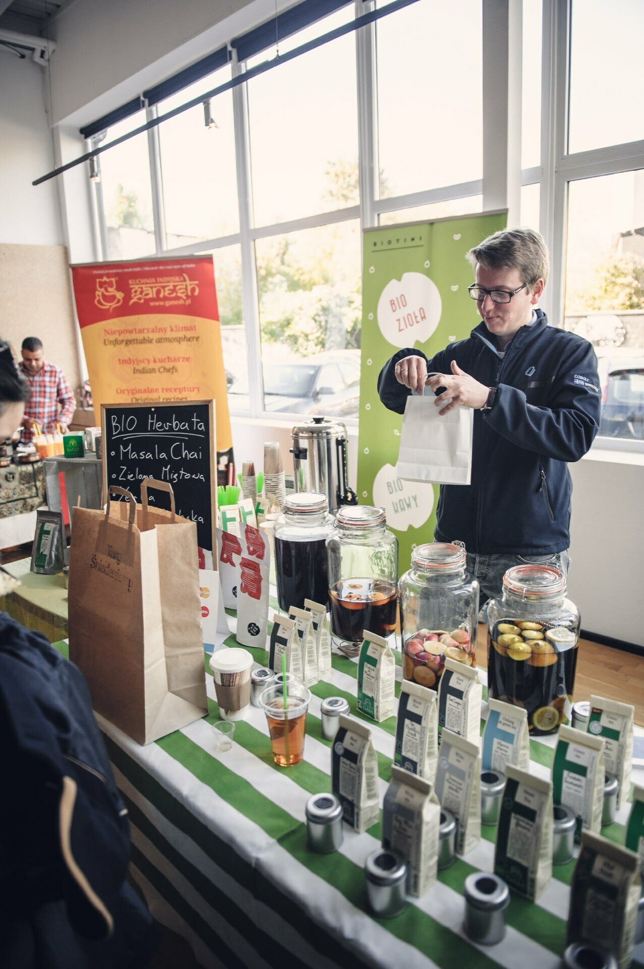 A vendor at a stall prepares a paper bag for a customer. The stall, which is part of a bustling culinary fair, displays a variety of organic tea products in jars and boxes. A board behind the vendor lists the types of teas, and a green sign promoting organic products is visible in the background.  