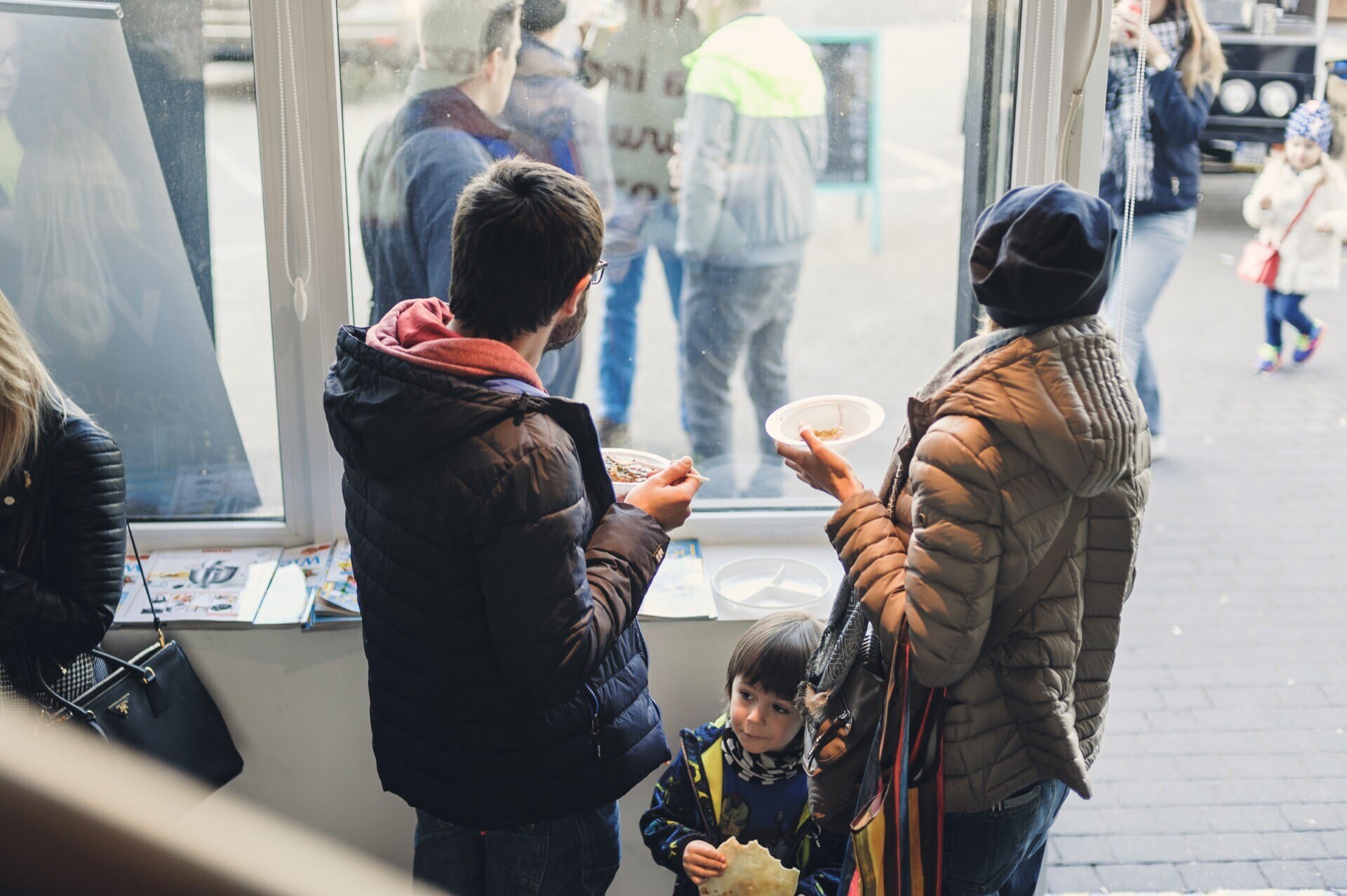 A family of three stands by the window eating. A child raises his head, holding his food, and the parents, immersed in conversation, hold their plates. Outside, several people can be seen walking or standing, as if at a food fair. The family is warmly dressed in jackets and hats.   