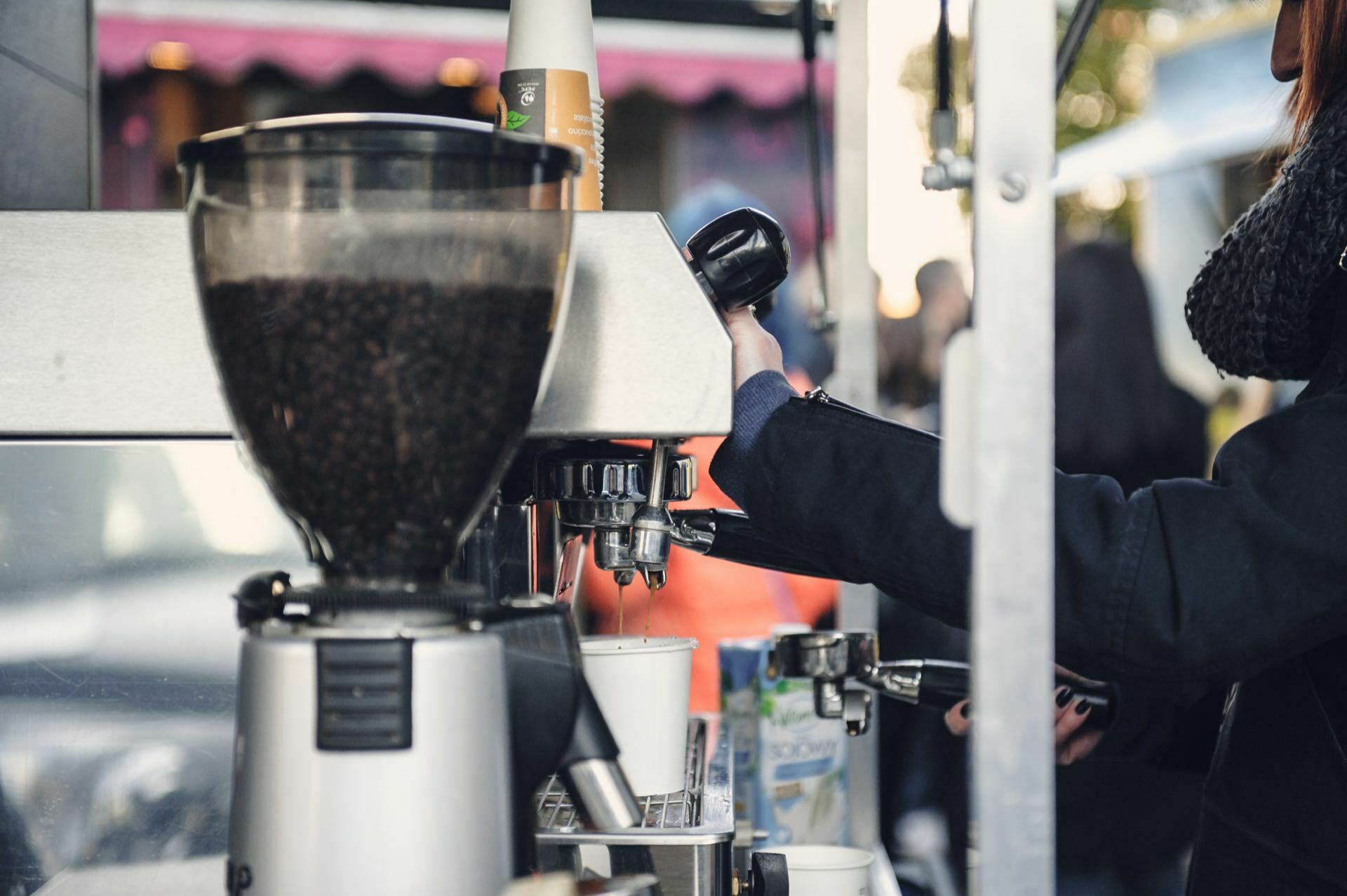 On the side, a person can be seen operating an espresso machine at an outdoor coffee stand during a culinary fair. Coffee beans can be seen in the grinder next to the machine, and paper cups are being filled with coffee. The person is wearing a dark coat and scarf. The blurred background suggests a street scene.   