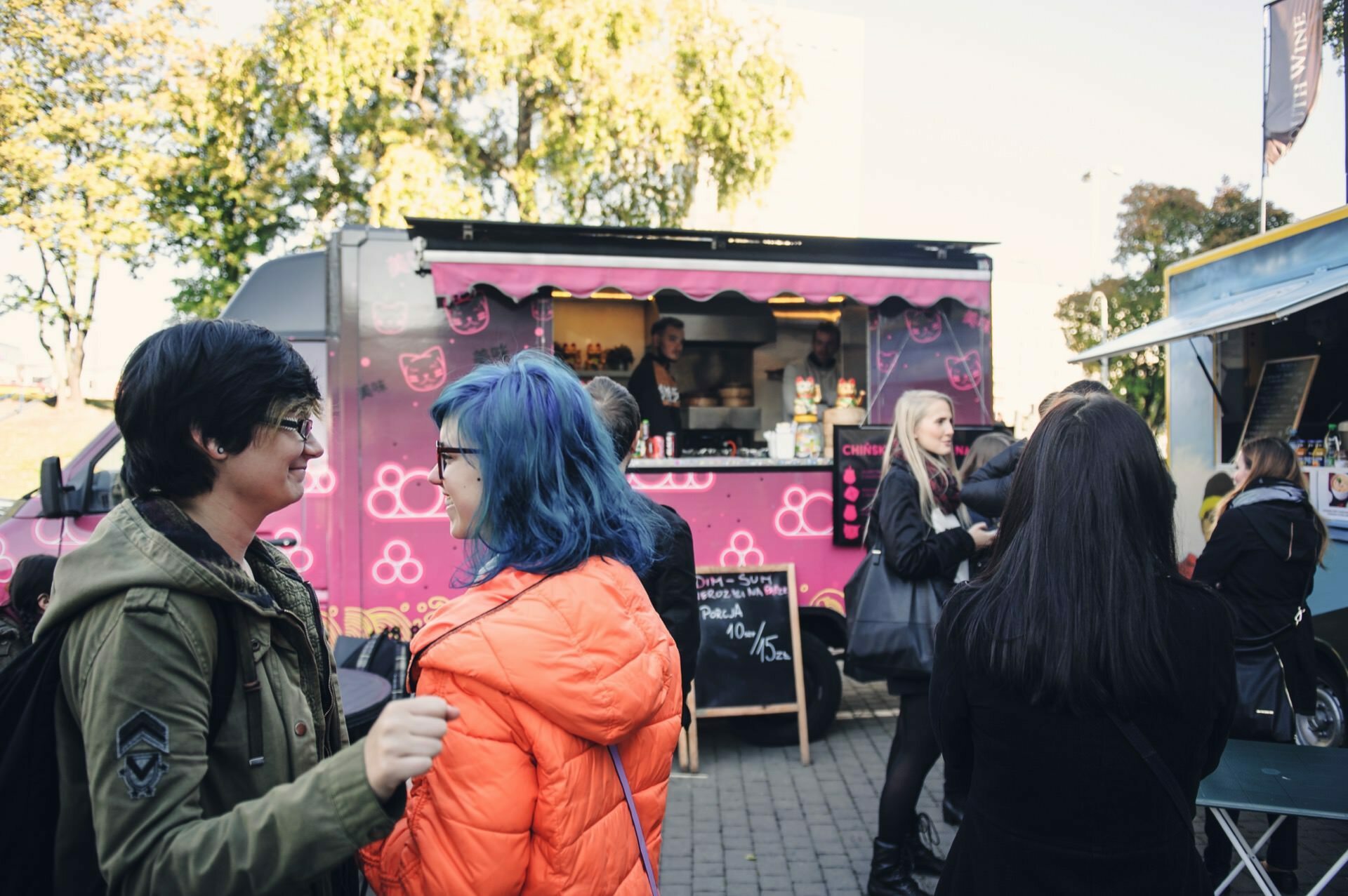 People gather in front of bustling food trucks at an open-air market resembling a food fair. One food truck is bright purple with cartoon images. In the foreground, two people in colorful jackets chat while others order from the food trucks.  