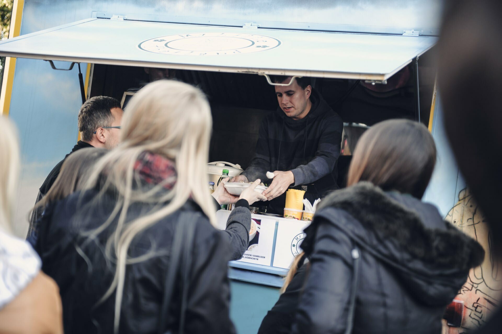 A vendor at a food truck serves a product to a customer. A small crowd of people waits in line in front of a blue truck with a menu on the side. The weather seems chilly as customers are dressed in jackets and scarves. It is reminiscent of scenes from bustling food markets.   