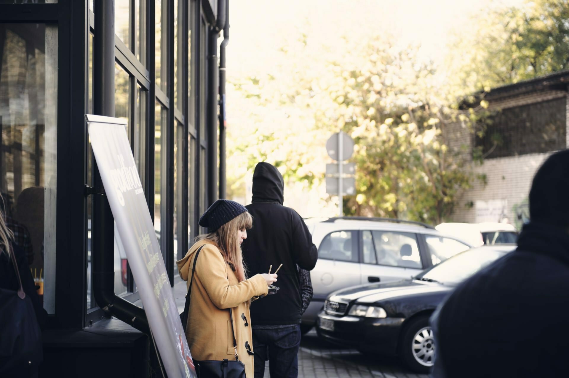 A person in a yellow coat and blue cap is looking at a phone, standing near a glass building. Another person in a black sweatshirt walks away, presumably heading toward the culinary fair. Several cars are parked along the street, and trees with autumn leaves can be seen in the background.  