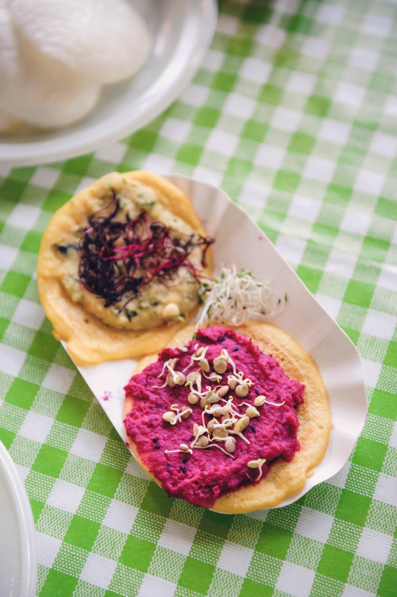 Two small tortillas with vibrant spreads lie on a paper tray on a green and white checkered tablecloth, reminiscent of a food fair. One tortilla has pink beet hummus and the other greenish cream, both decorated with microgreens. 