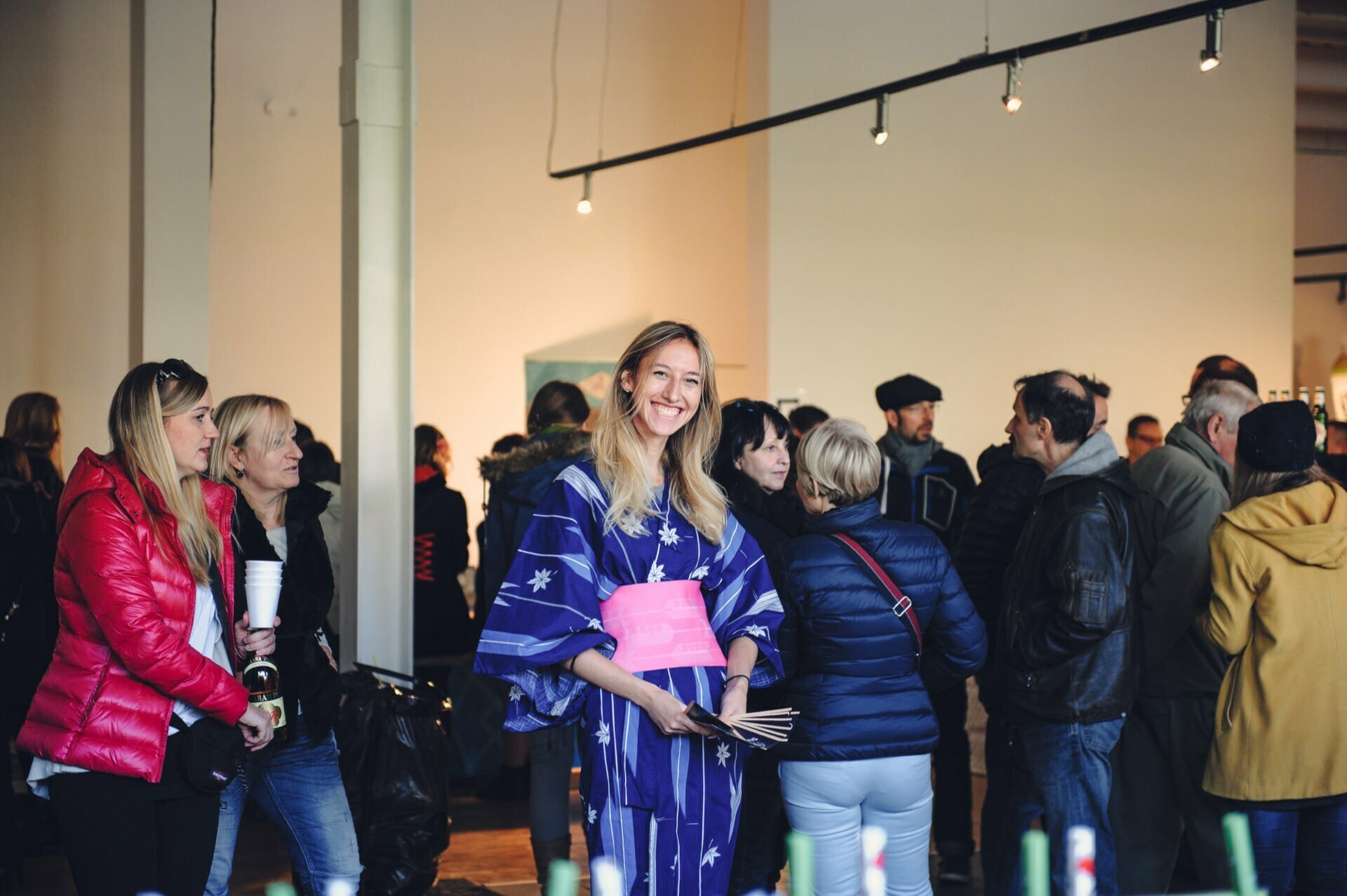 A woman dressed in a bright blue kimono with a pink obi smiles at the camera while standing in a crowded room. The background is filled with people talking, some wearing jackets and holding drinks, creating a casual, festive atmosphere reminiscent of a food fair. 