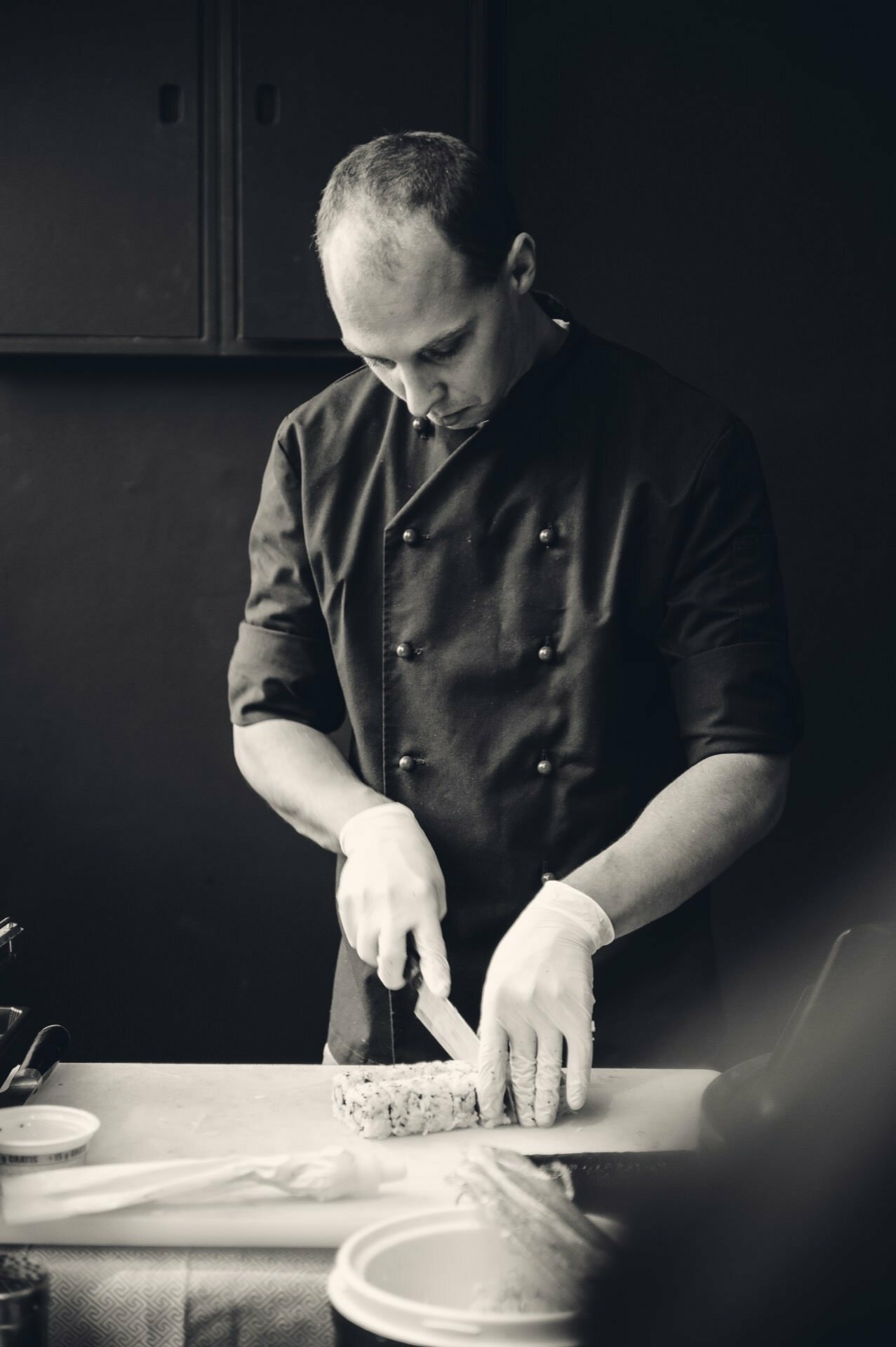 The chef, wearing a dark double-breasted coat and gloves, carefully slices food on a cutting board. The environment resembles a professional kitchen, perhaps at a culinary fair. The black and white image focuses on his precise cutting technique.  