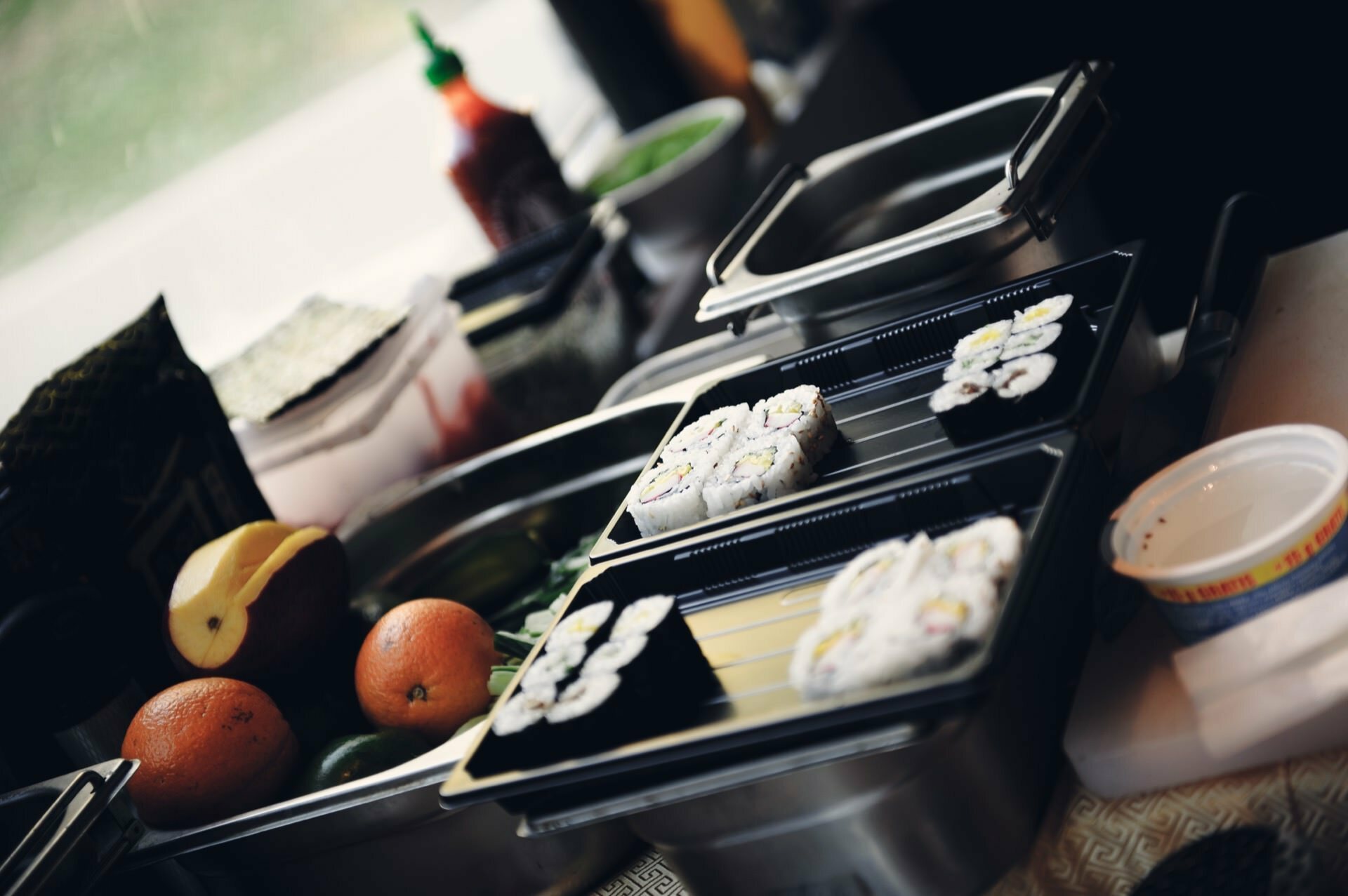 Close-up of a kitchen countertop filled with various ingredients for preparing sushi, reminiscent of a culinary fair. Two trays of sushi rolls are prominently displayed, each filled with carefully cut pieces. Fresh fruit, sauces and cooking utensils are visible in the background.  