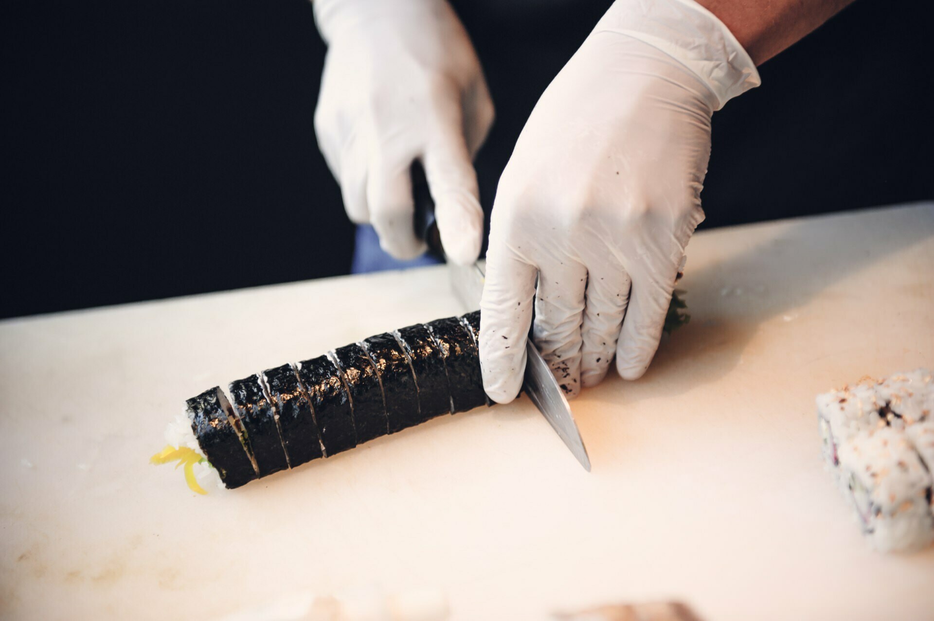 At a food fair, a person wearing white gloves cuts a sushi roll with a sharp knife on a white cutting board. The sushi roll wrapped in seaweed and filled with visible ingredients shows culinary precision. In the background, another sushi roll can be seen partially out of frame.  